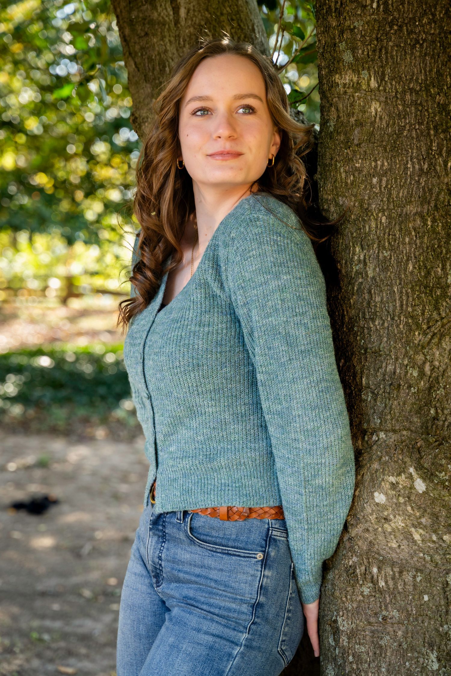 Young woman in a teal cardigan and jeans leaning against a tree in a sunlit park.