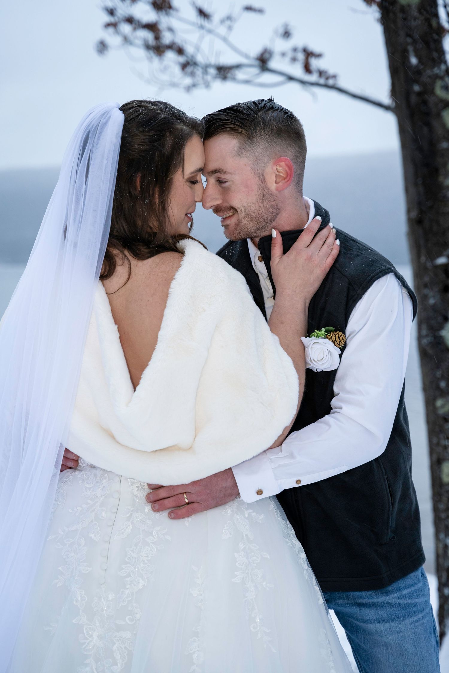 Bride in white lace gown with fur wrap and groom in vest share an intimate forehead touch in winter.