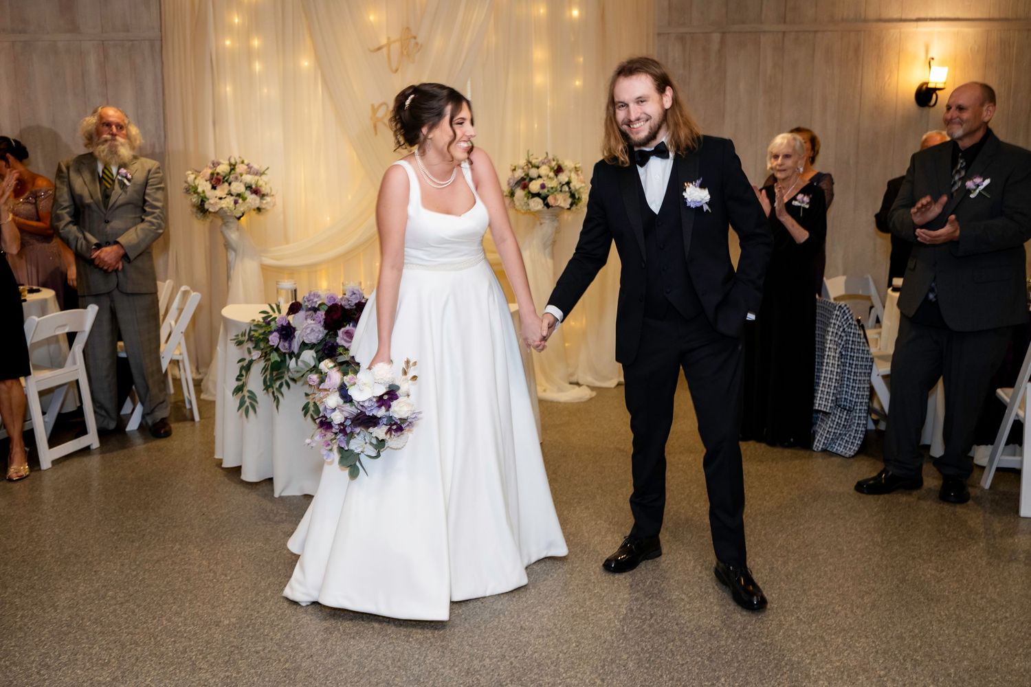 Bride carrying purple bouquet and long-haired groom enter wedding reception hall hand in hand.