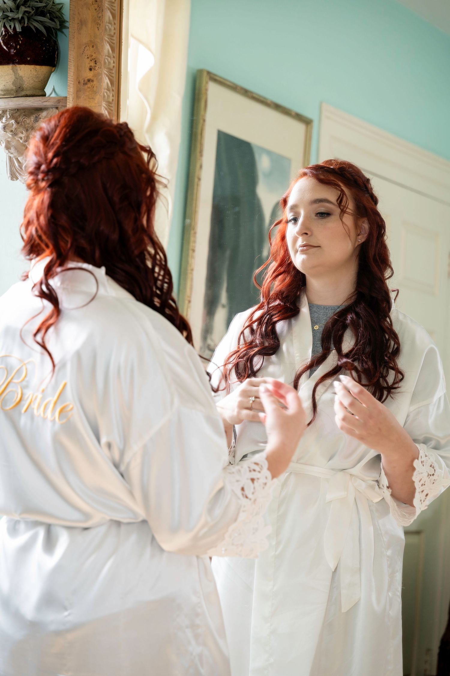 Red-haired bride in white satin Bride robe admires herself in mirror during wedding morning preparations.