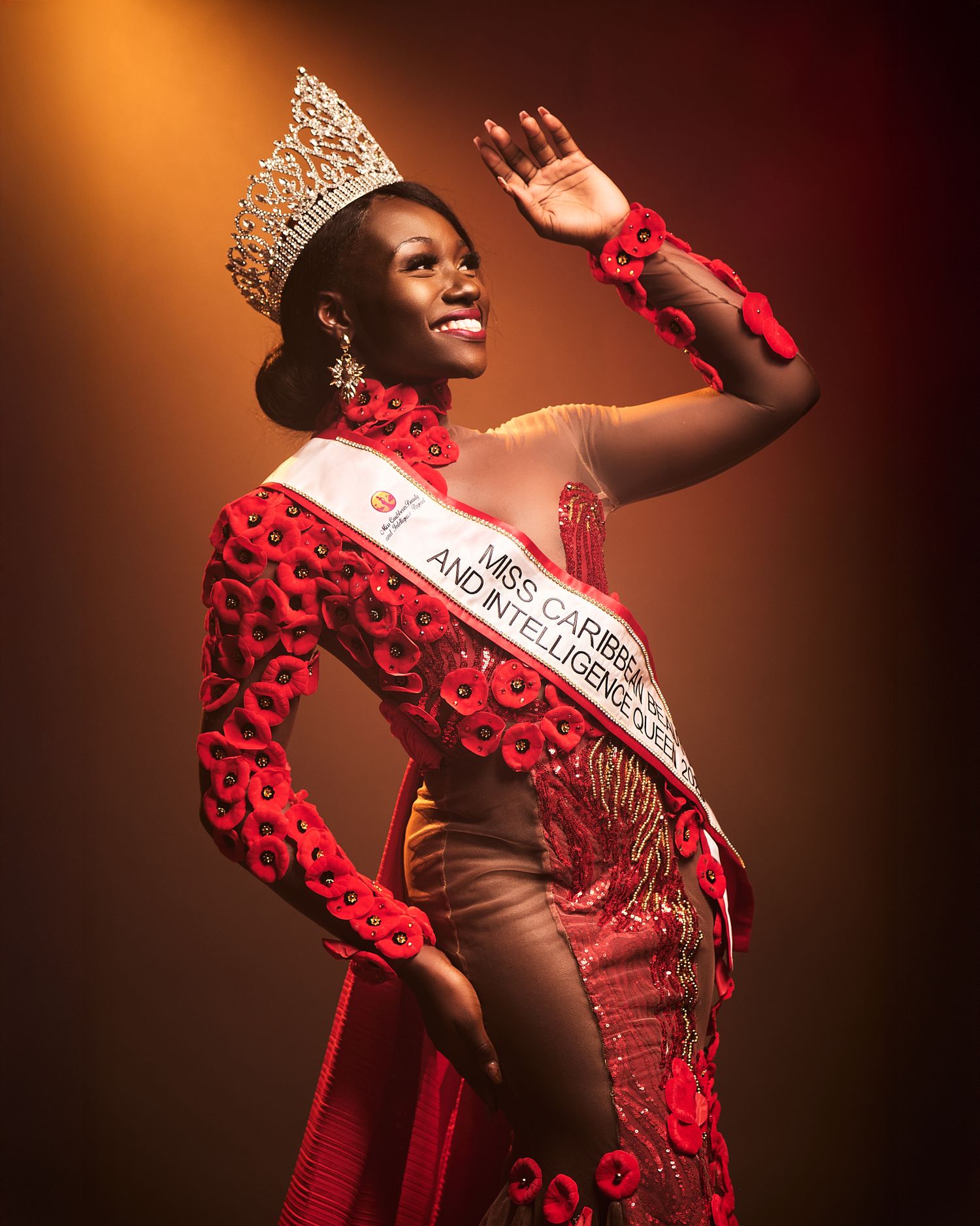 Miss Caribbean beauty queen in red gown and crown waves gracefully on stage under warm spotlight.