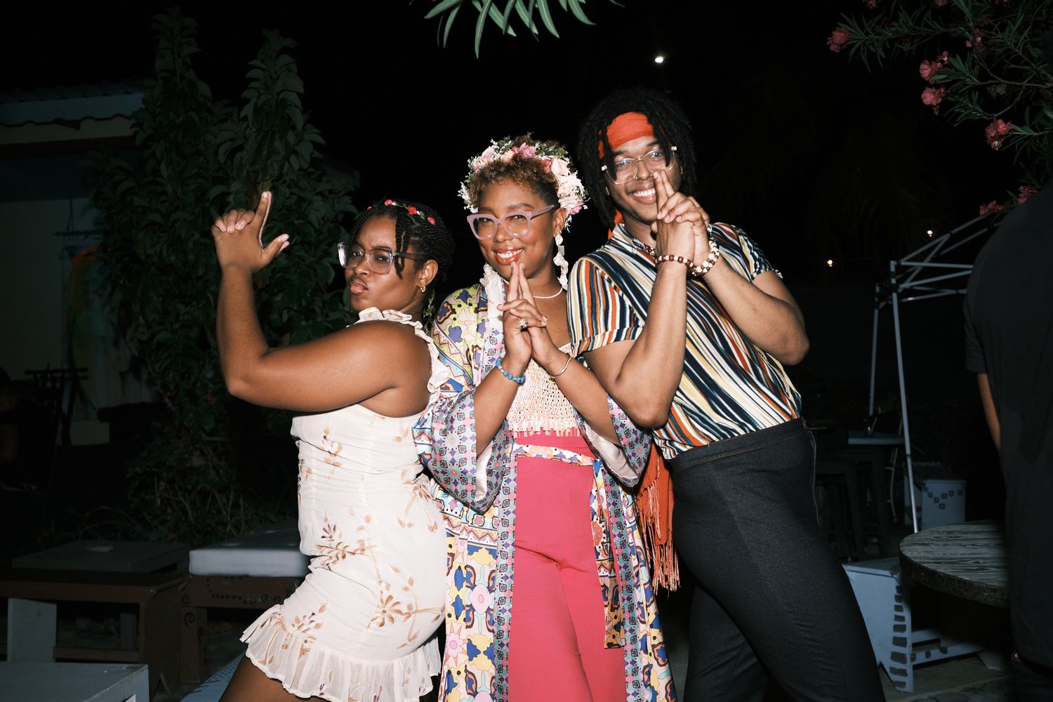 Three friends posing and smiling at a festive outdoor night party, wearing colorful outfits and floral accessories.