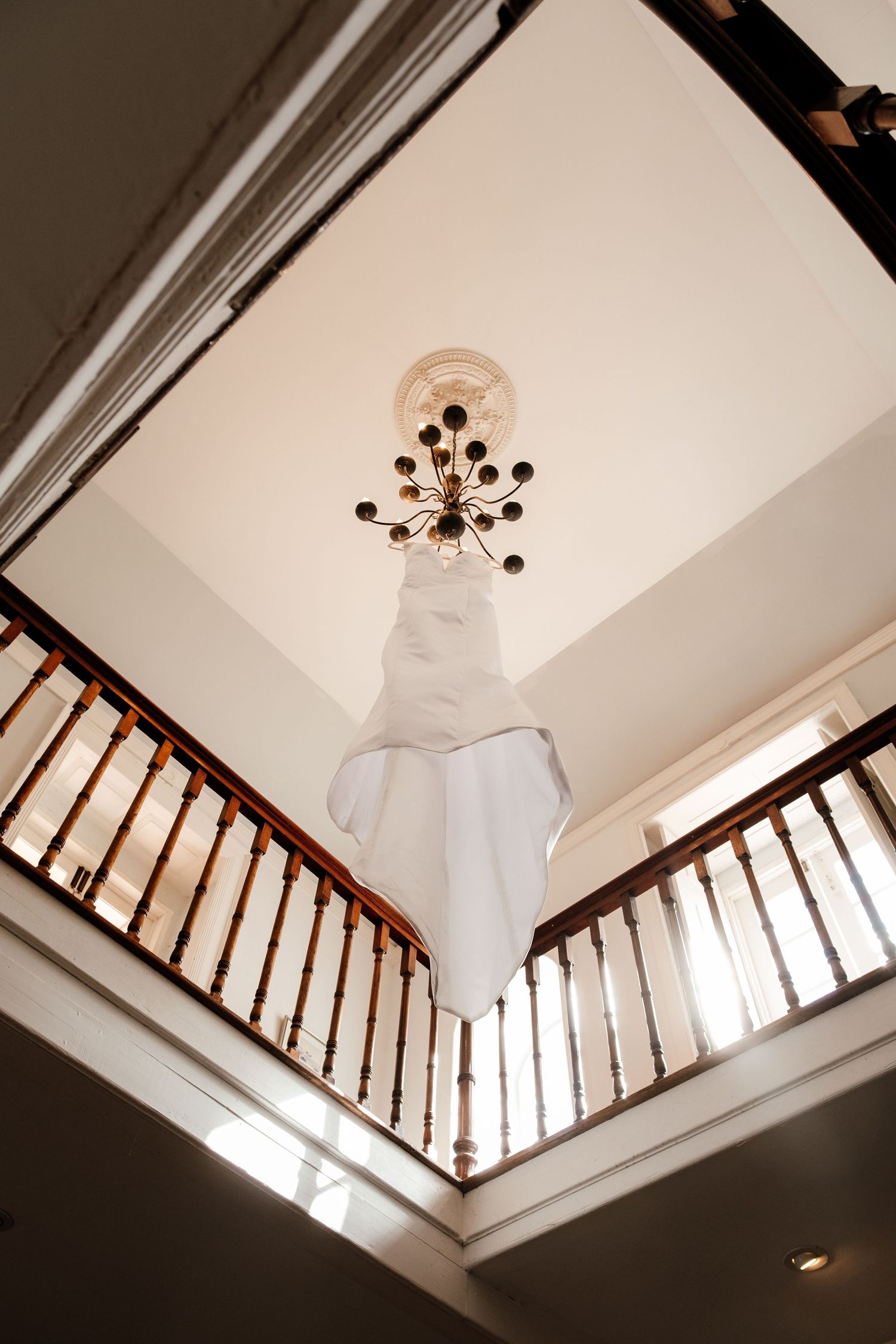 White wedding dress hanging from chandelier in elegant two-story foyer with wooden staircase railing.
