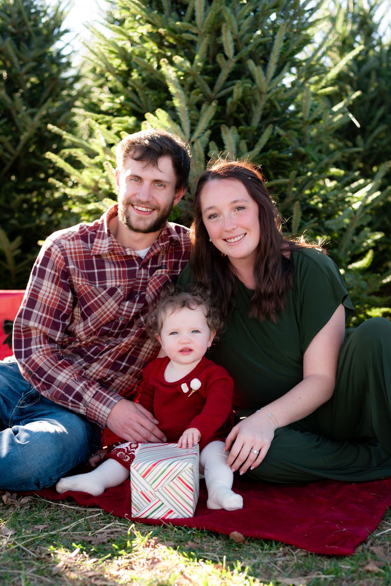 Family relaxes on red blanket during outdoor Christmas photo session near evergreen trees.