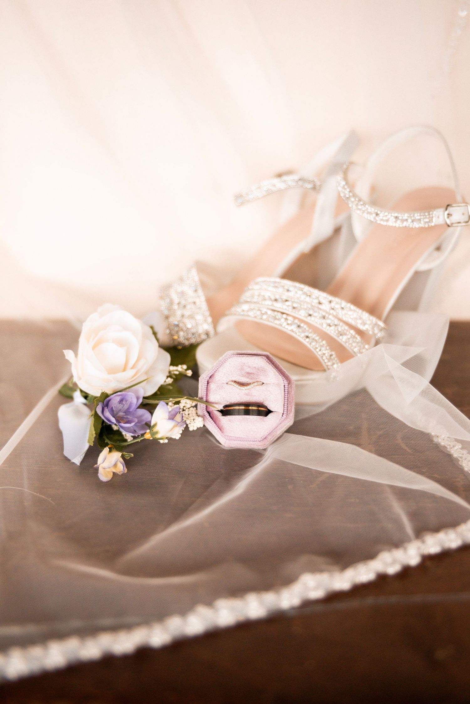 Elegant bridal flat lay featuring pearl heels, pink ring box with wedding bands, and floral boutonniere on a lace veil.