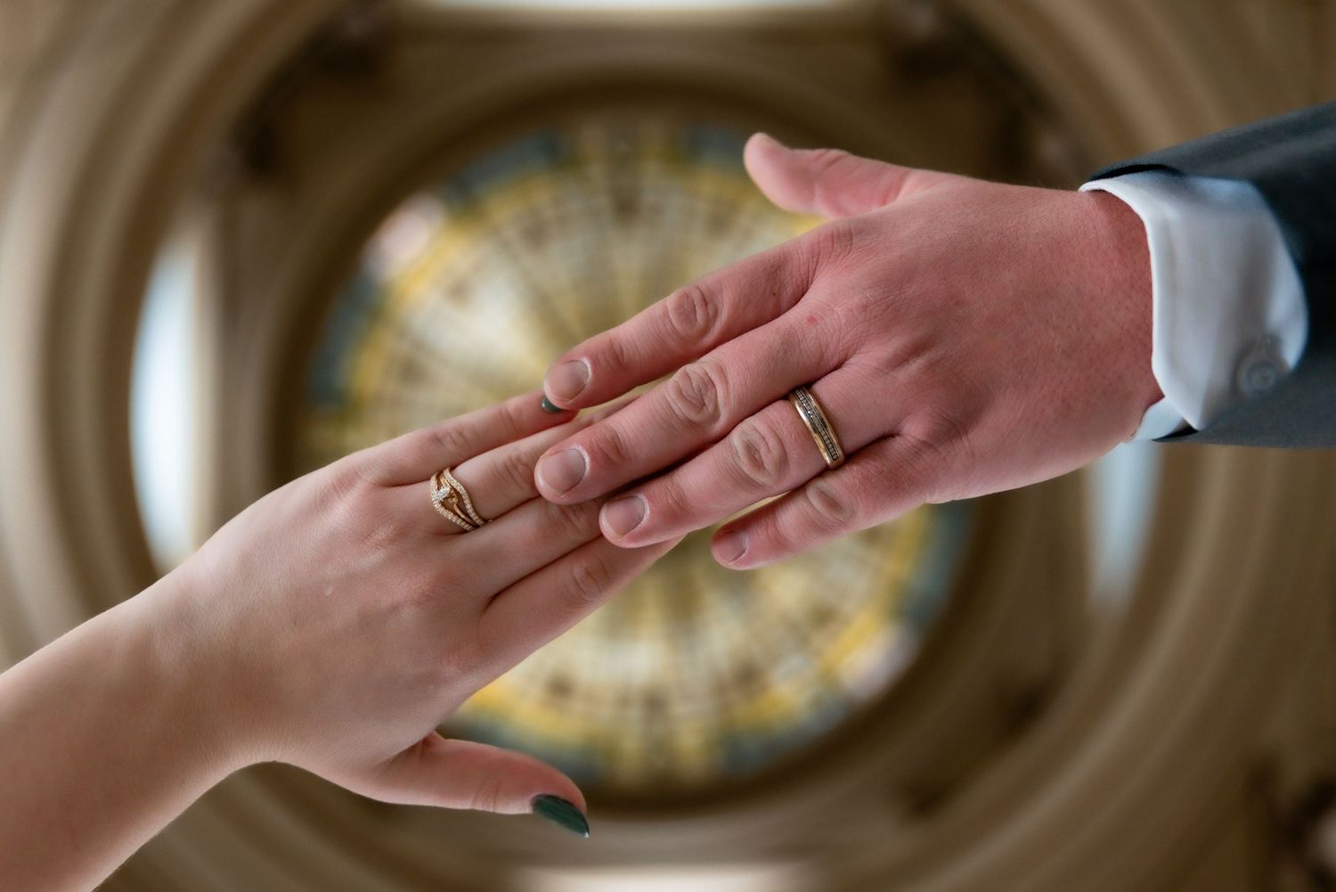 Two hands touching, adorned with gold wedding rings, with a blurred stained glass ceiling in the background.