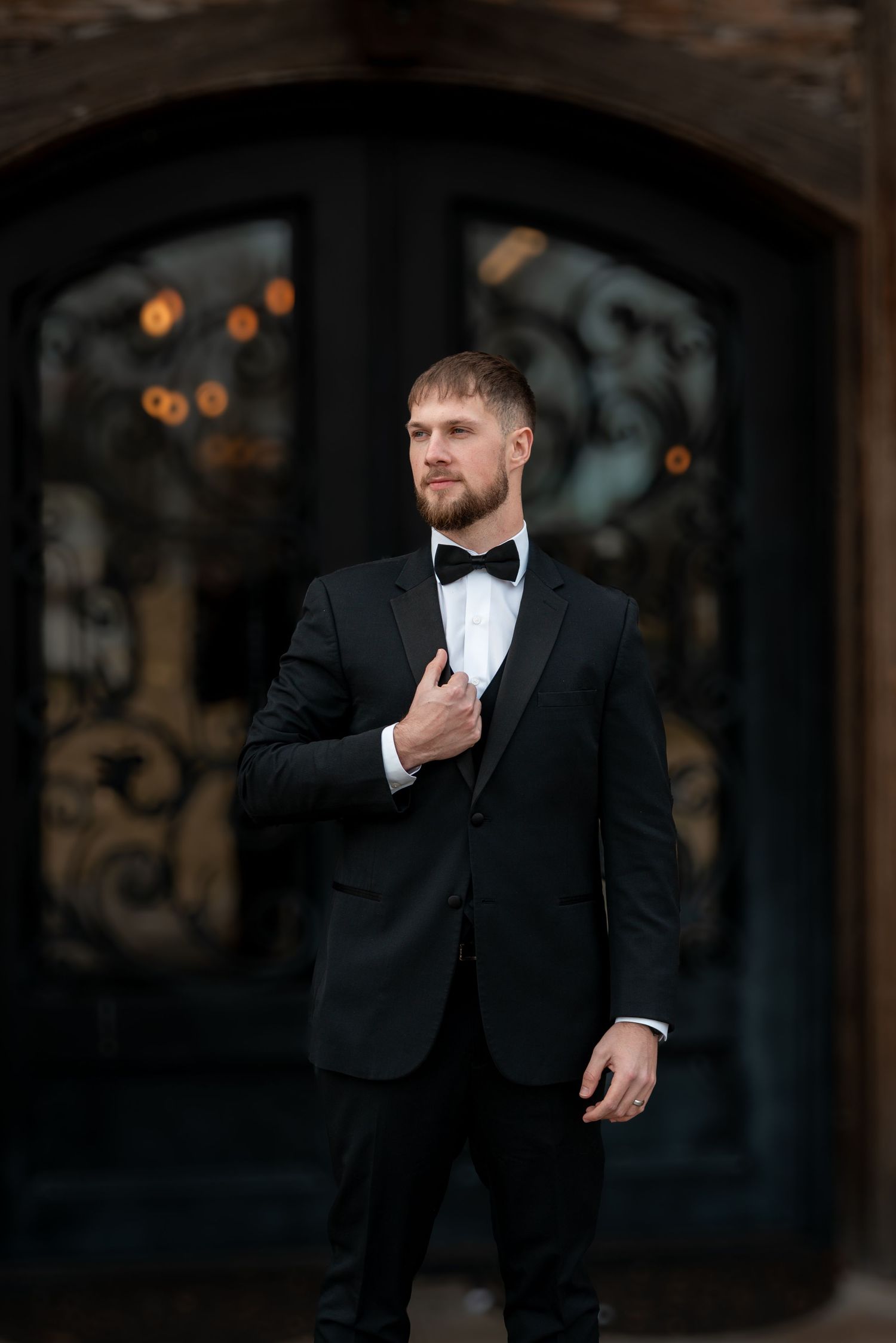 Groom in classic black tuxedo poses in front of ornate iron doors.