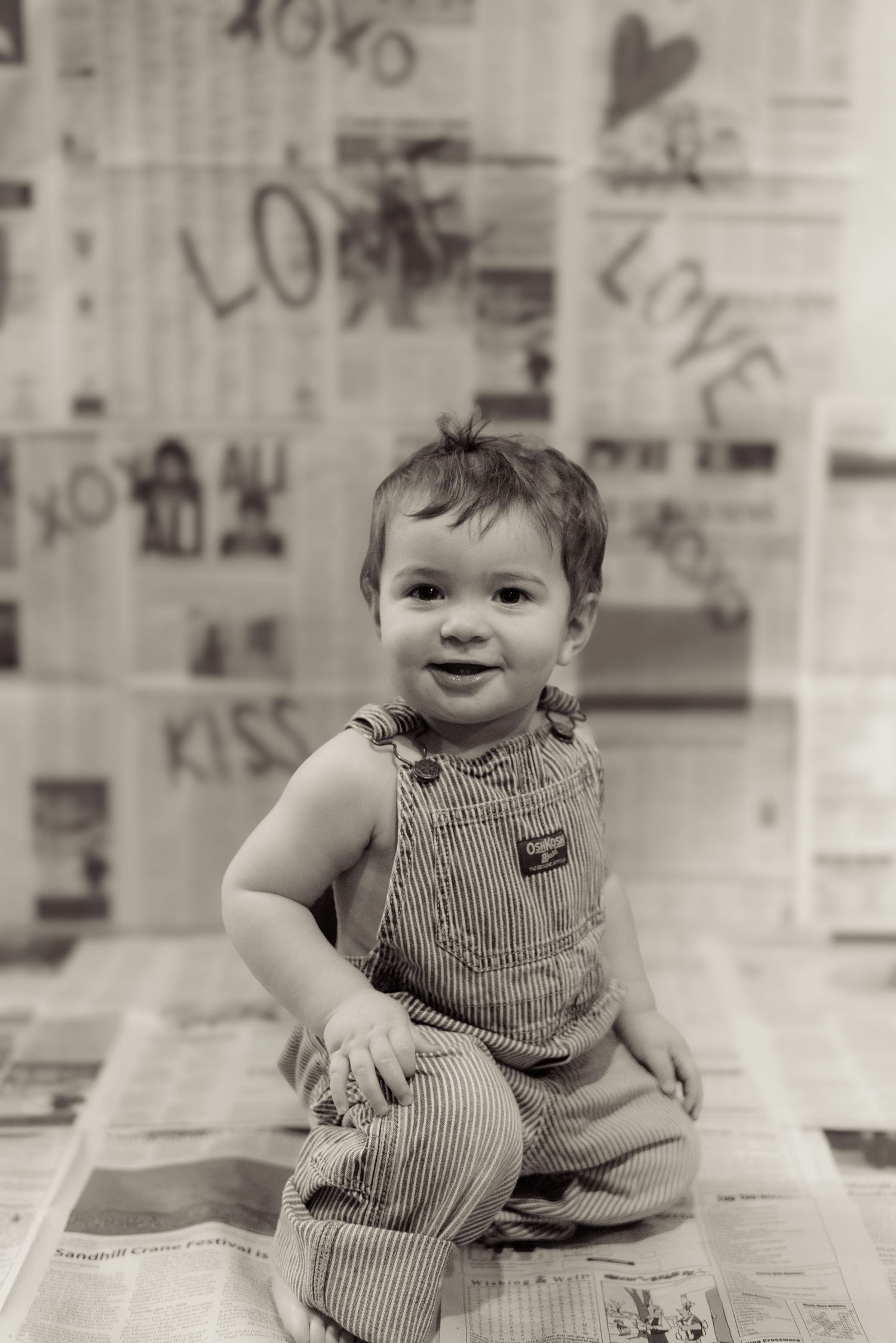 A vintage-style photo of a toddler in striped overalls sitting on newspaper.