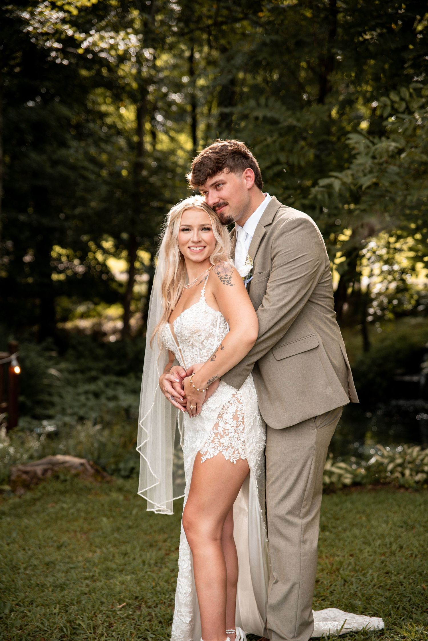 Bride in lace mini dress with veil and groom in tan suit embrace outdoors in a lush garden wedding portrait.