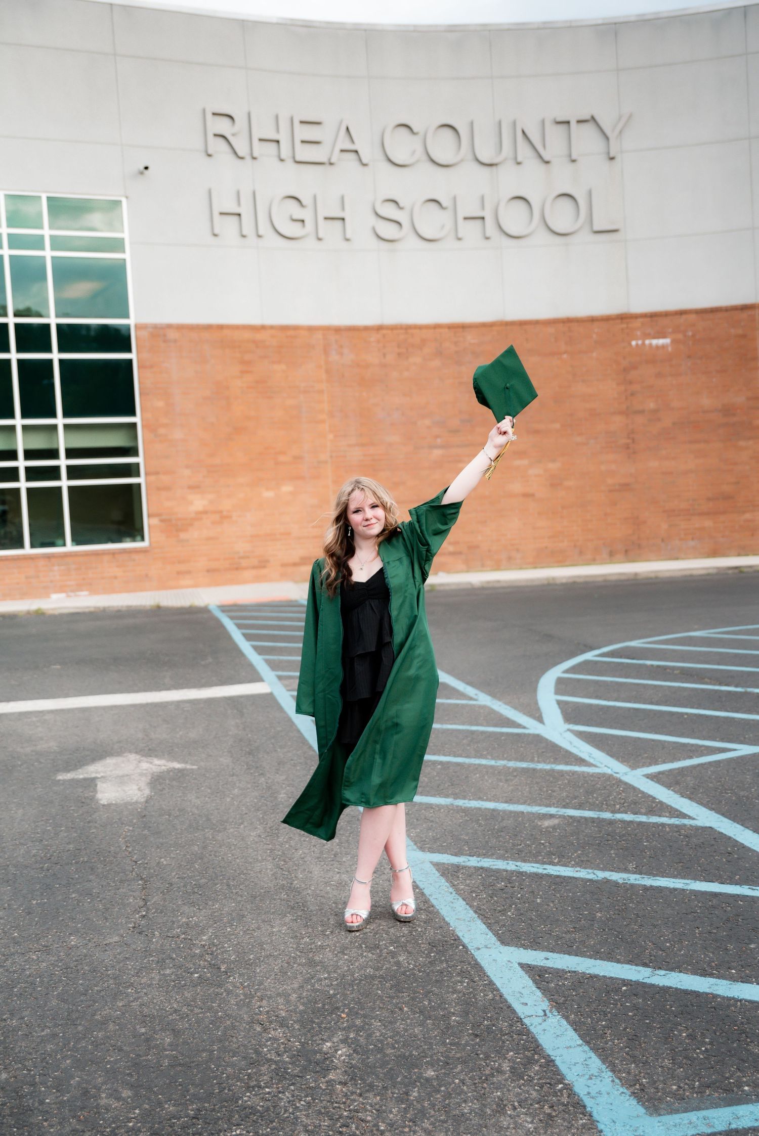 Graduate in green cap and gown raises hat in front of Rhea County High School.