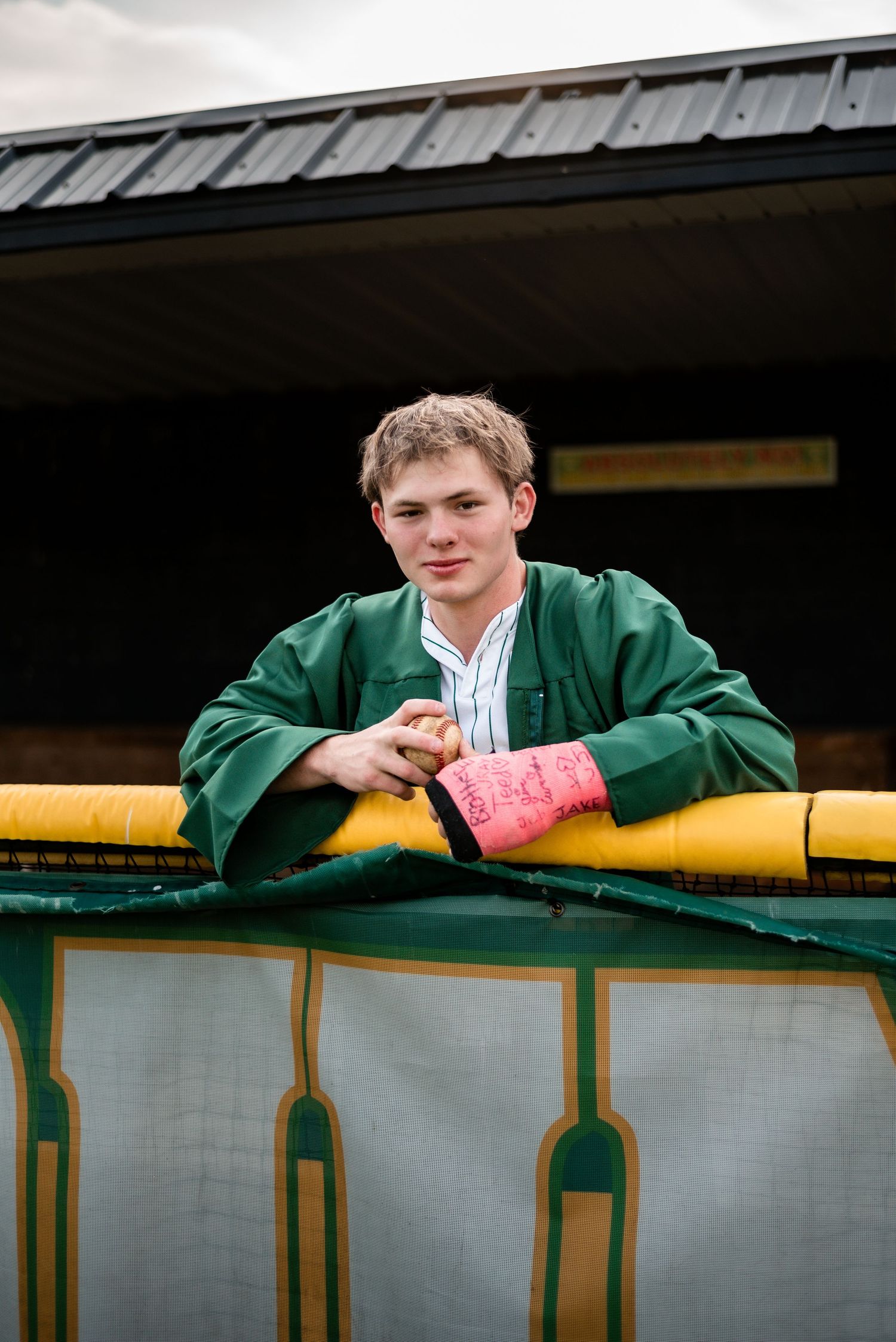 Young man in green graduation gown with pink wrist wrap leaning on yellow railing outdoors.