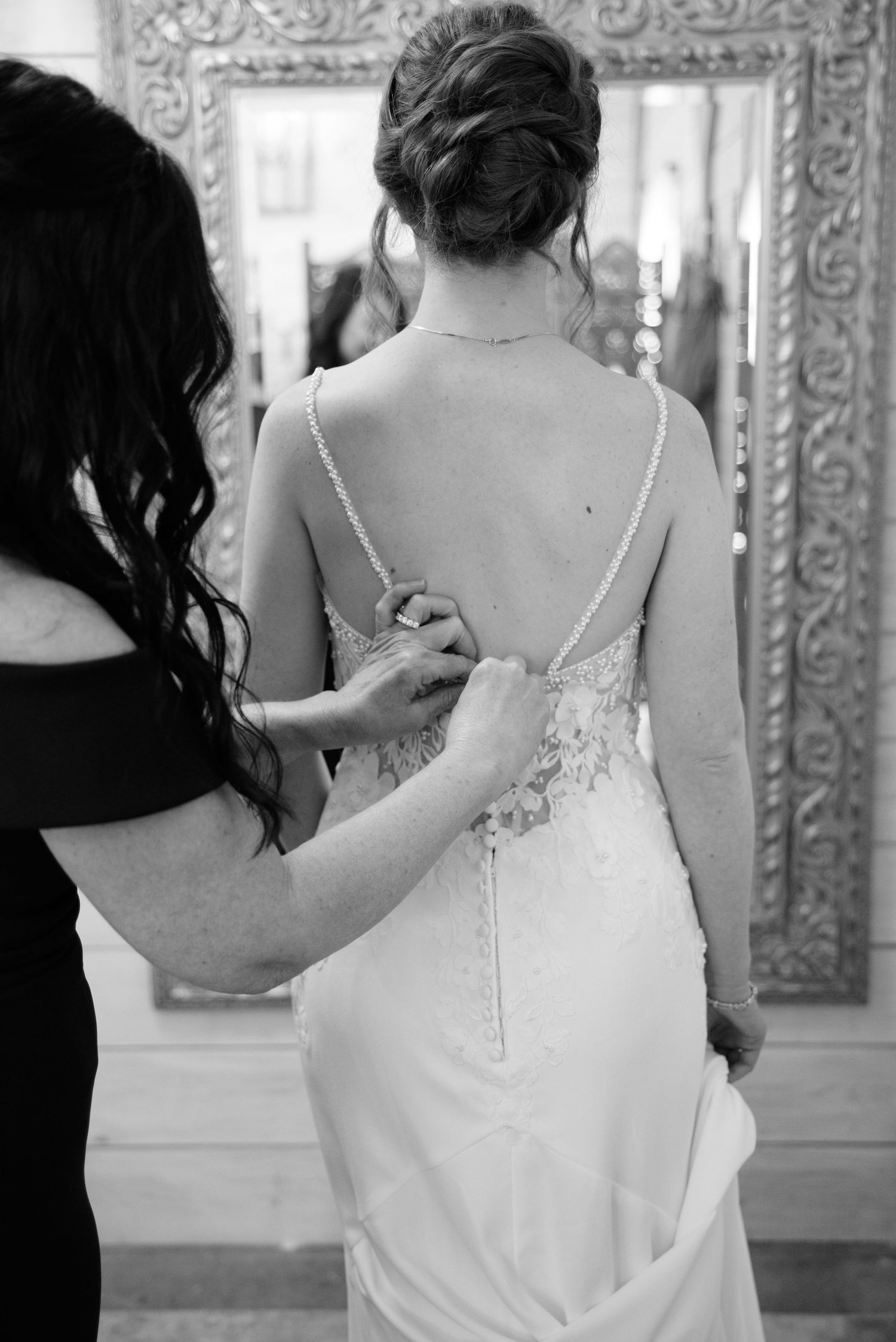 Black and white photo of a bride having her lace wedding dress buttoned up by a helper before her ceremony.