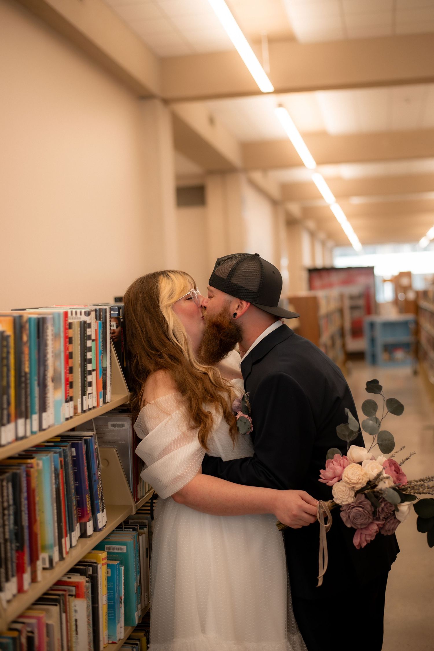 A couple shares a romantic kiss in a library aisle, bride in white gown holding floral bouquet, groom in black cap.