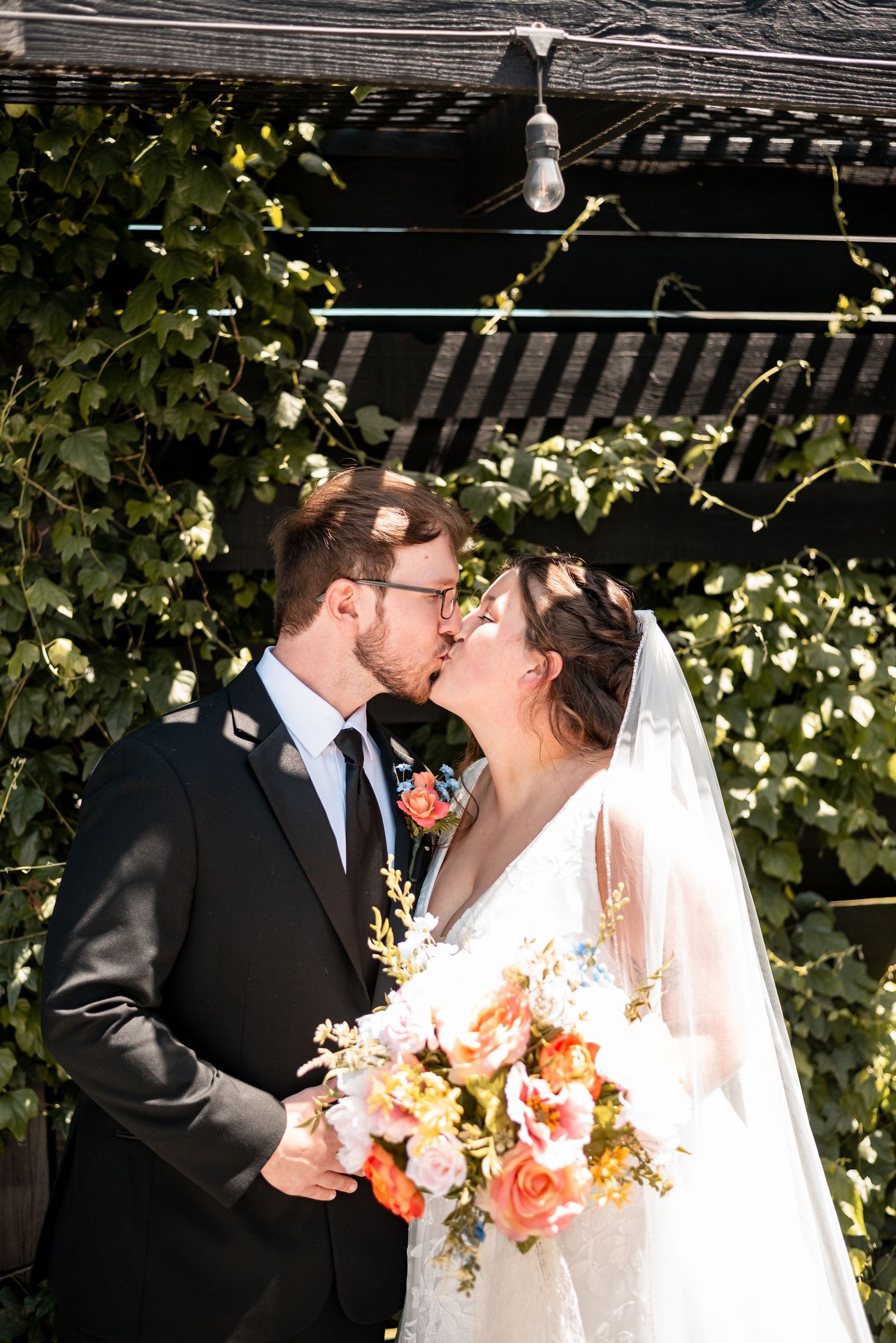 Bride and groom share a kiss outdoors, bride holding a colorful bouquet, surrounded by lush green ivy.
