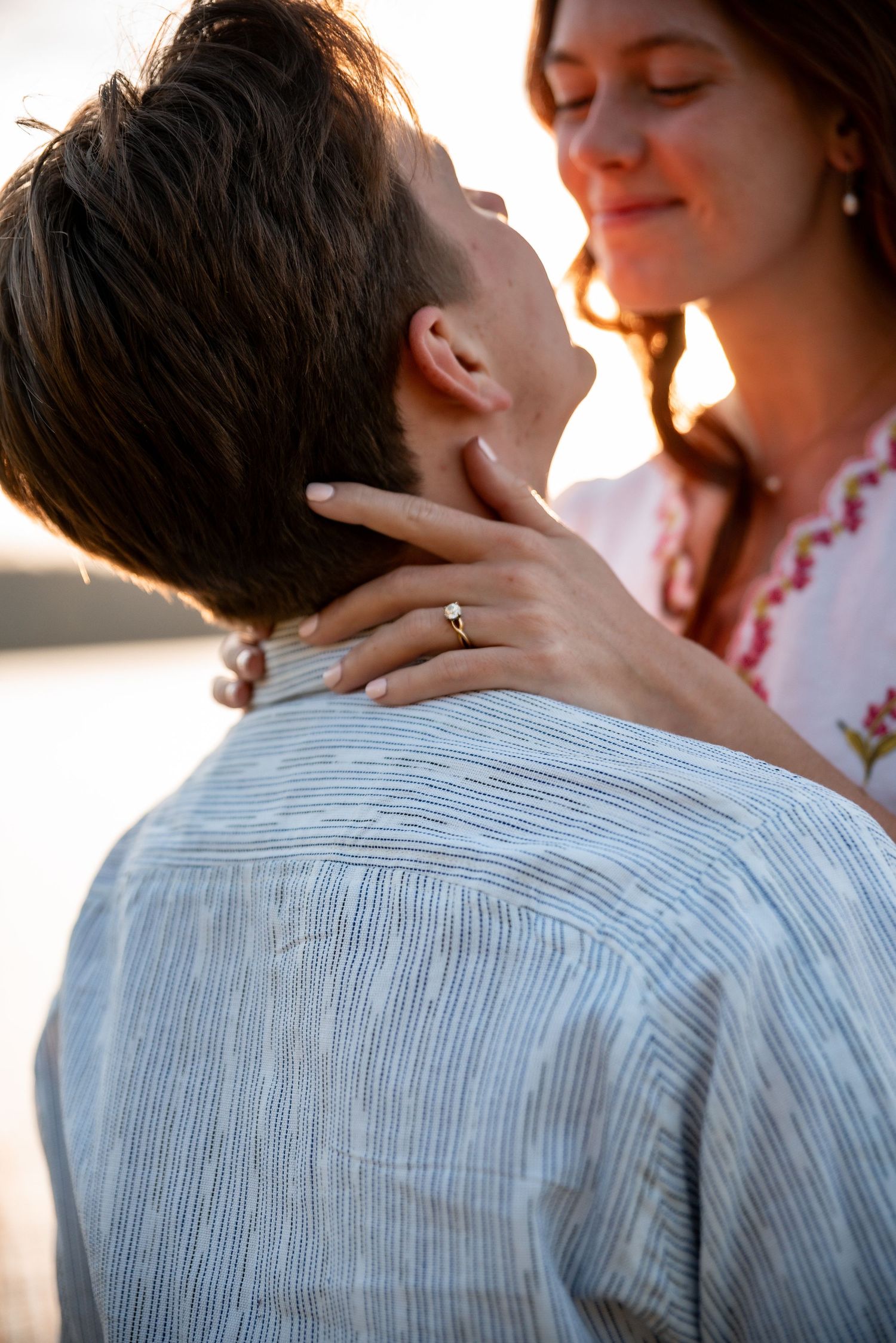Couple embracing at sunset, woman smiling with engagement ring visible on hand resting on man's neck.
