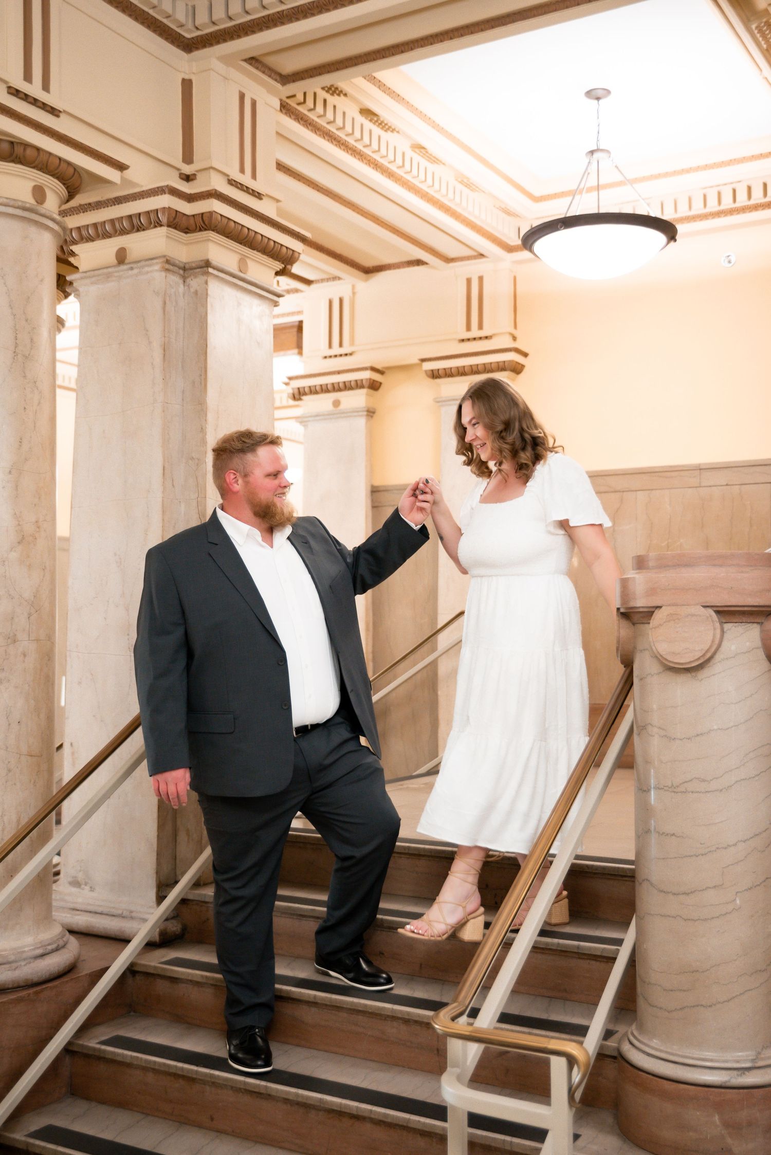 A couple holds hands on grand marble stairs, man in dark suit, woman in white dress, in an elegant building interior.