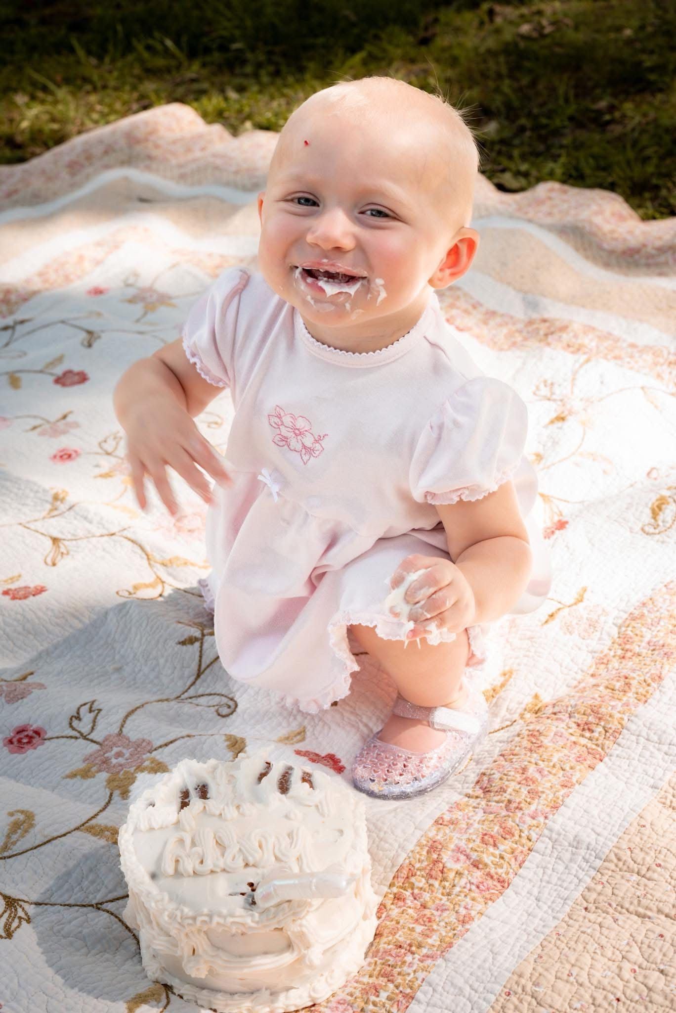 Baby in white dress sits on floral blanket outdoors.