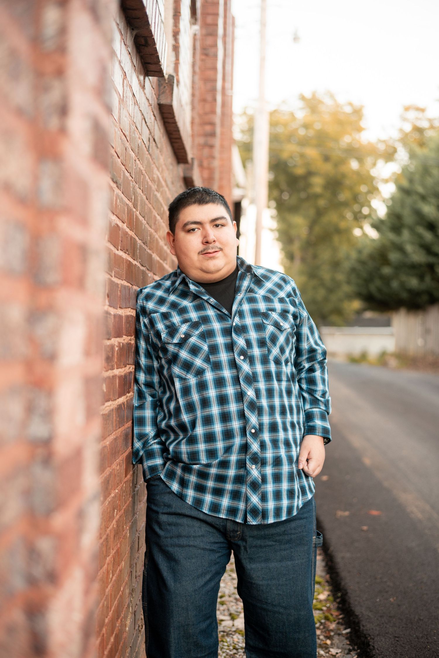Person in plaid shirt poses against brick wall during outdoor portrait session.