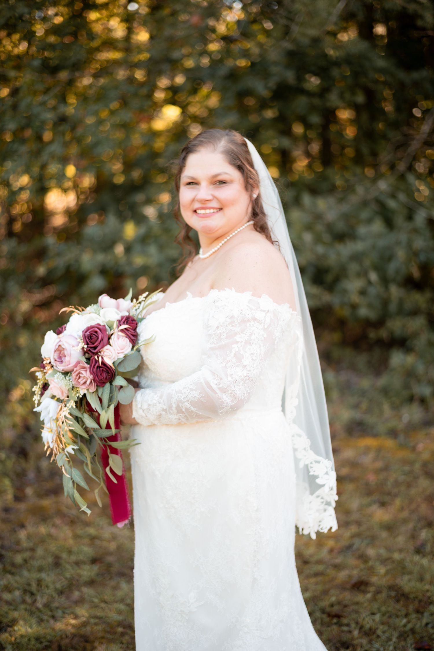 Beautiful bridal portrait in a white lace dress with burgundy and pink bouquet against autumn foliage.