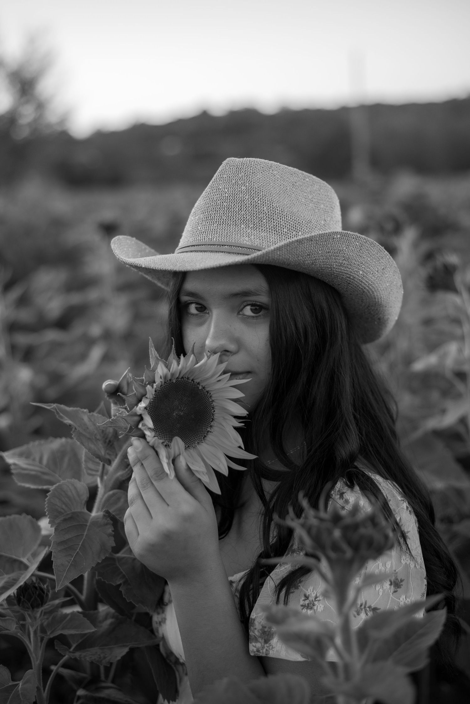 A person in a cowboy hat poses artistically with sunflowers in black and white photography.