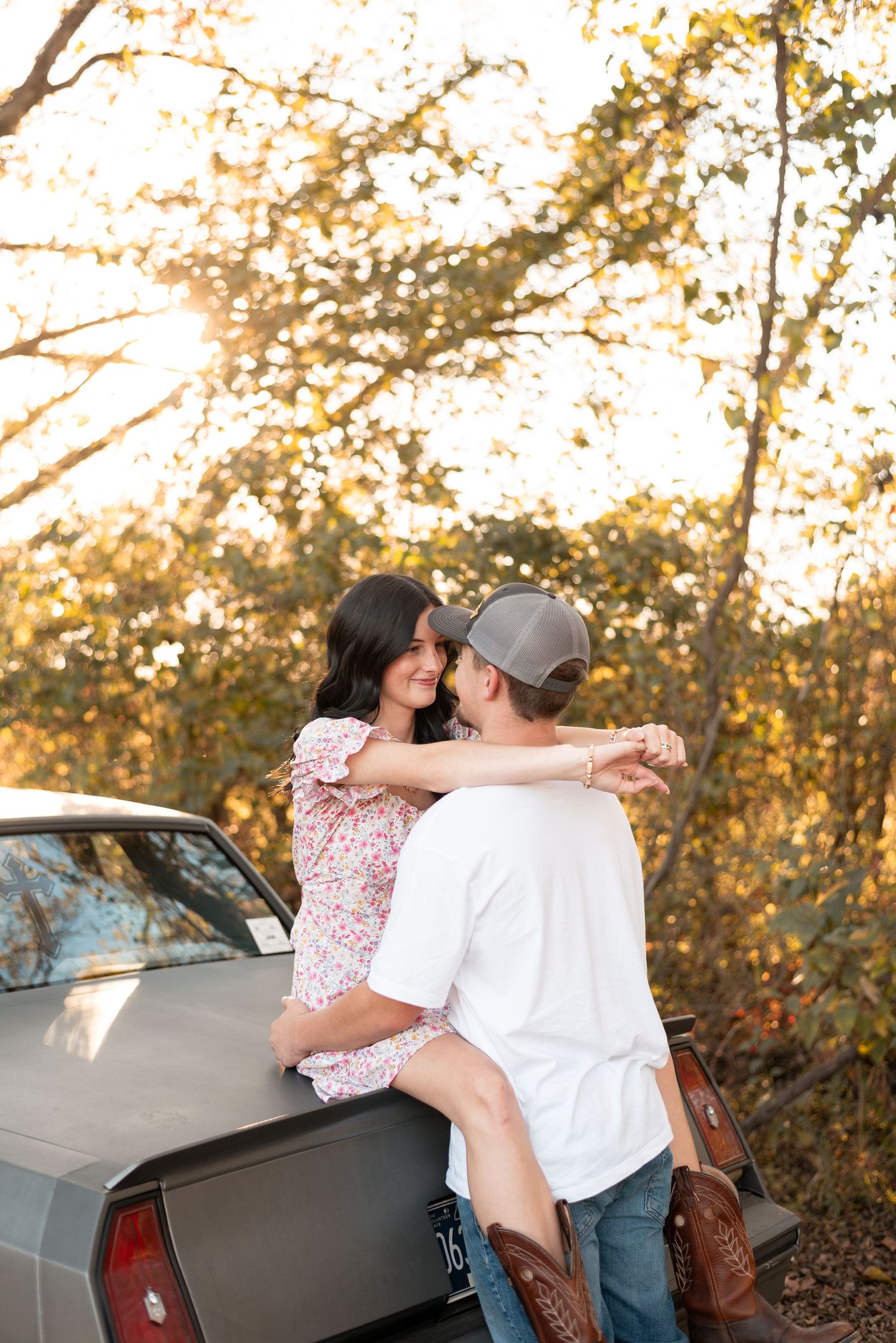 A couple embraces near a car during golden hour with autumn foliage in the background.