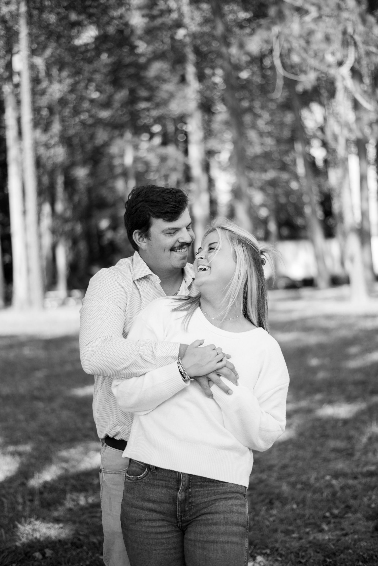 A couple embraces while laughing together in a wooded outdoor setting.