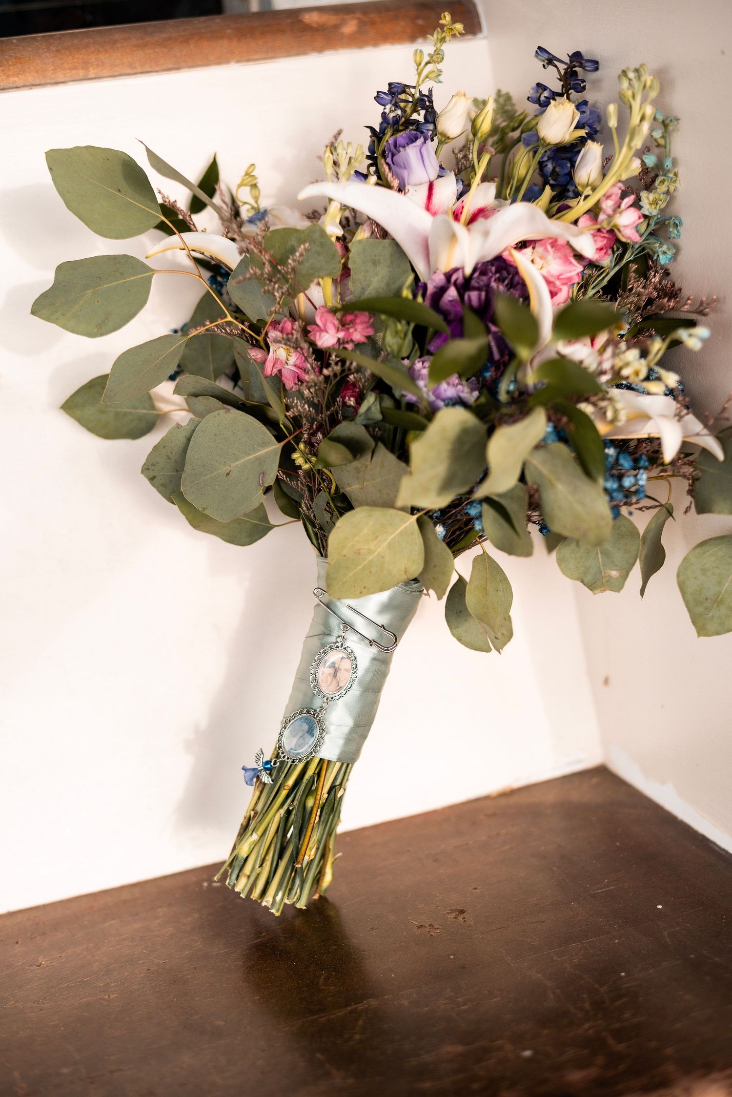 Colorful wedding bouquet with eucalyptus, pink roses, purple flowers, wrapped in light blue ribbon, resting on wooden surface.