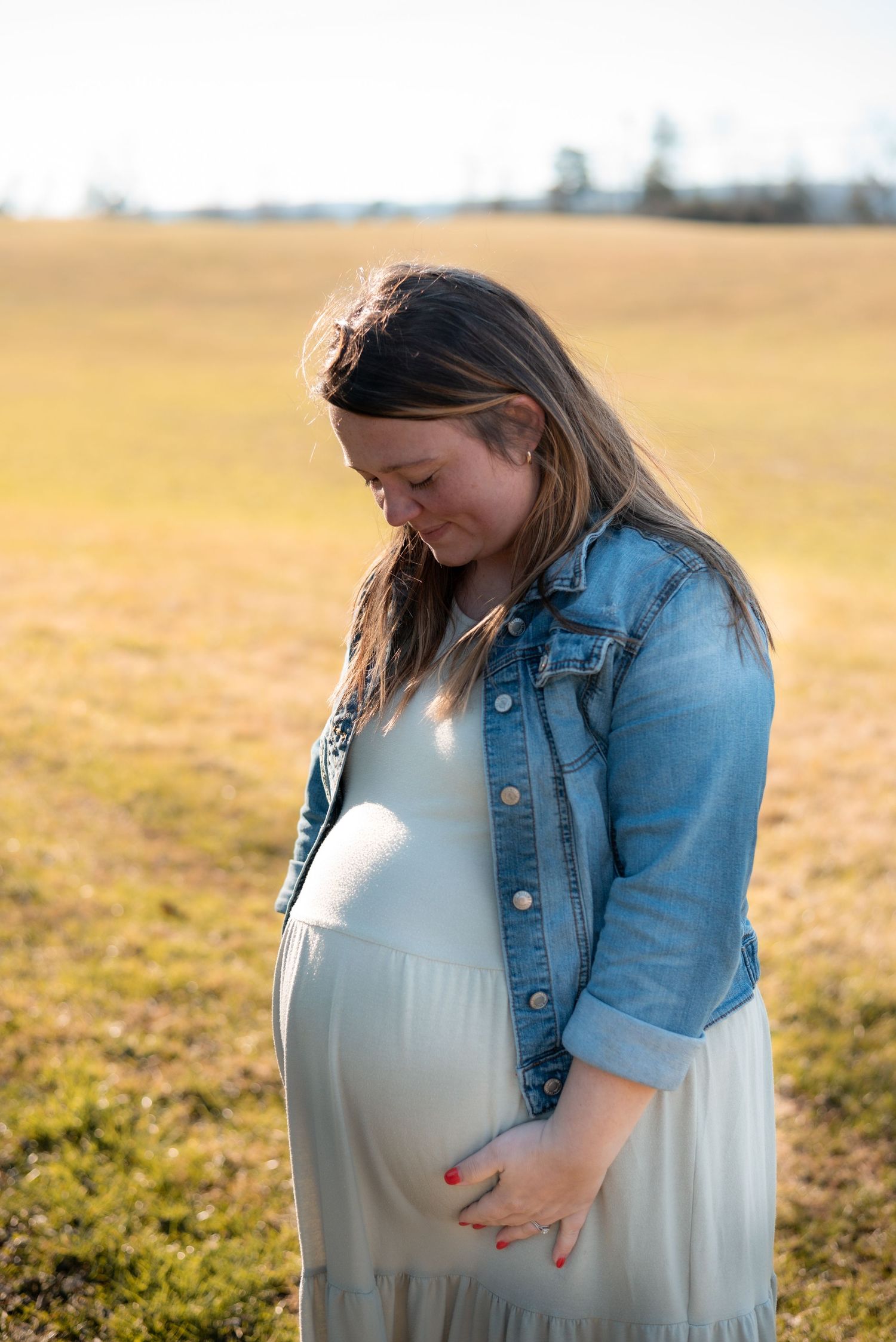 Expectant mother in a white dress and denim jacket touches her baby bump in a golden field.