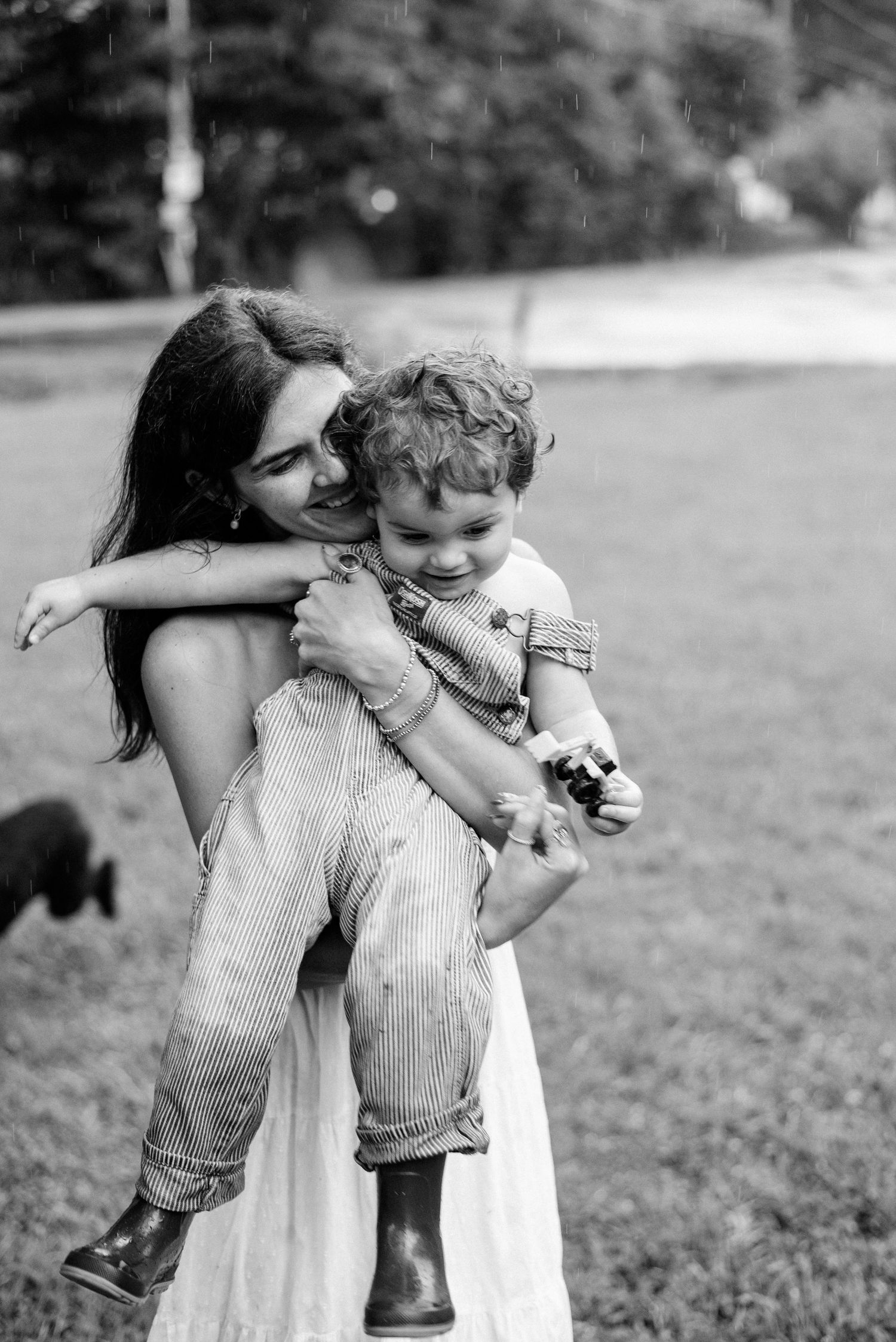 Black and white photo of a smiling mother hugging and lifting her young toddler outdoors on a grassy lawn.