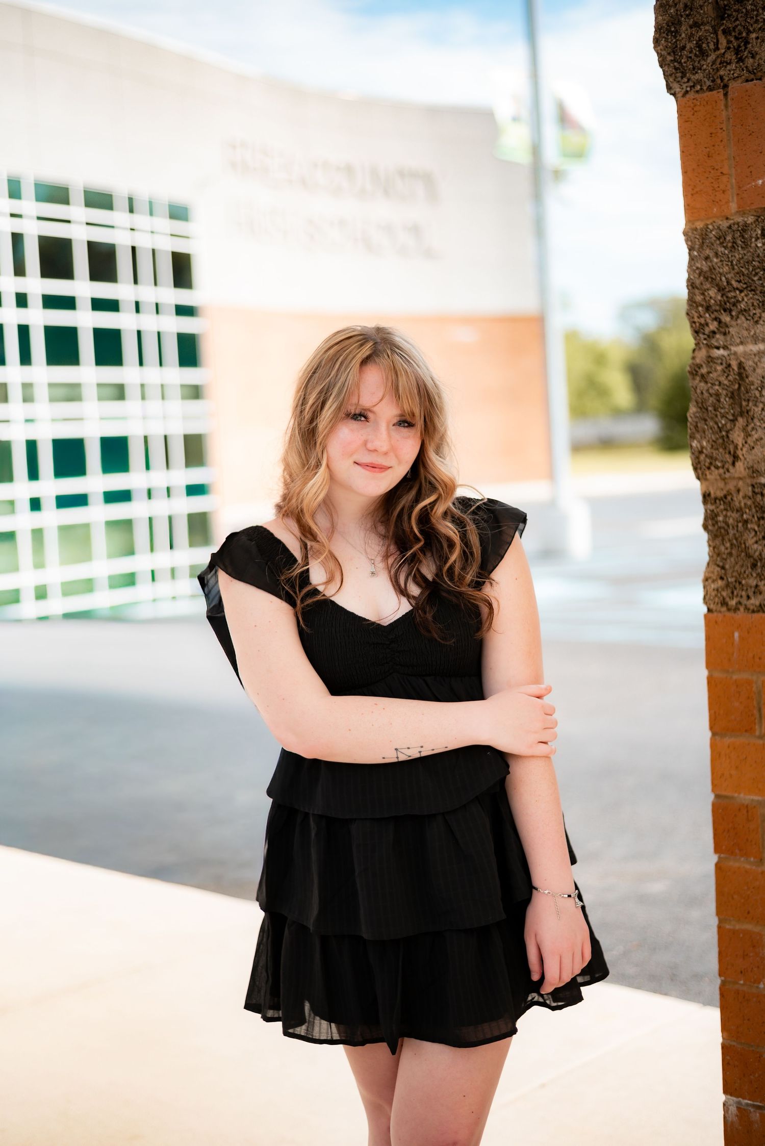 Young woman in black ruffled dress poses by brick pillar outside high school.