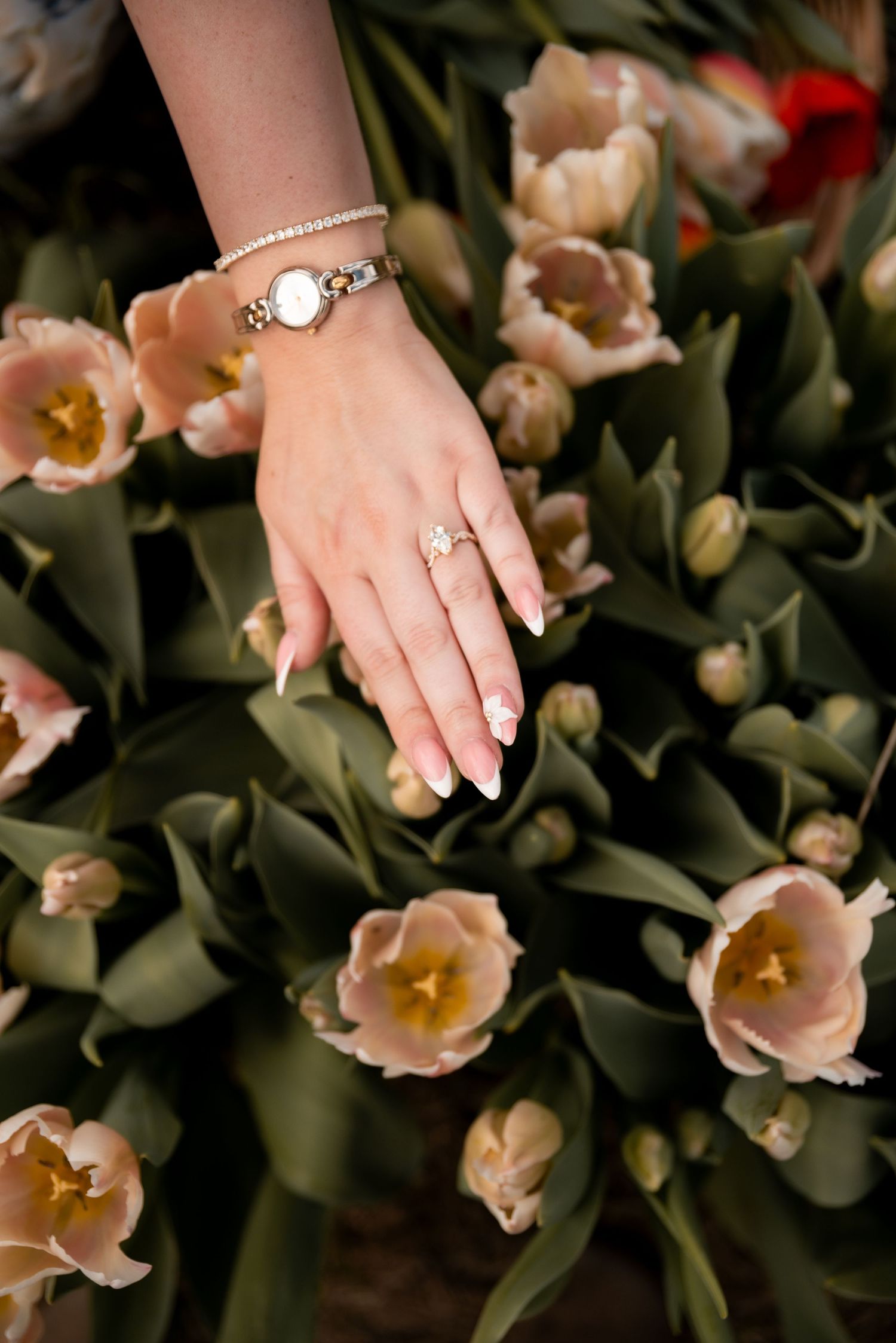 A woman's hand adorned with a gold watch, bracelet, and floral ring rests among blooming pink tulips.