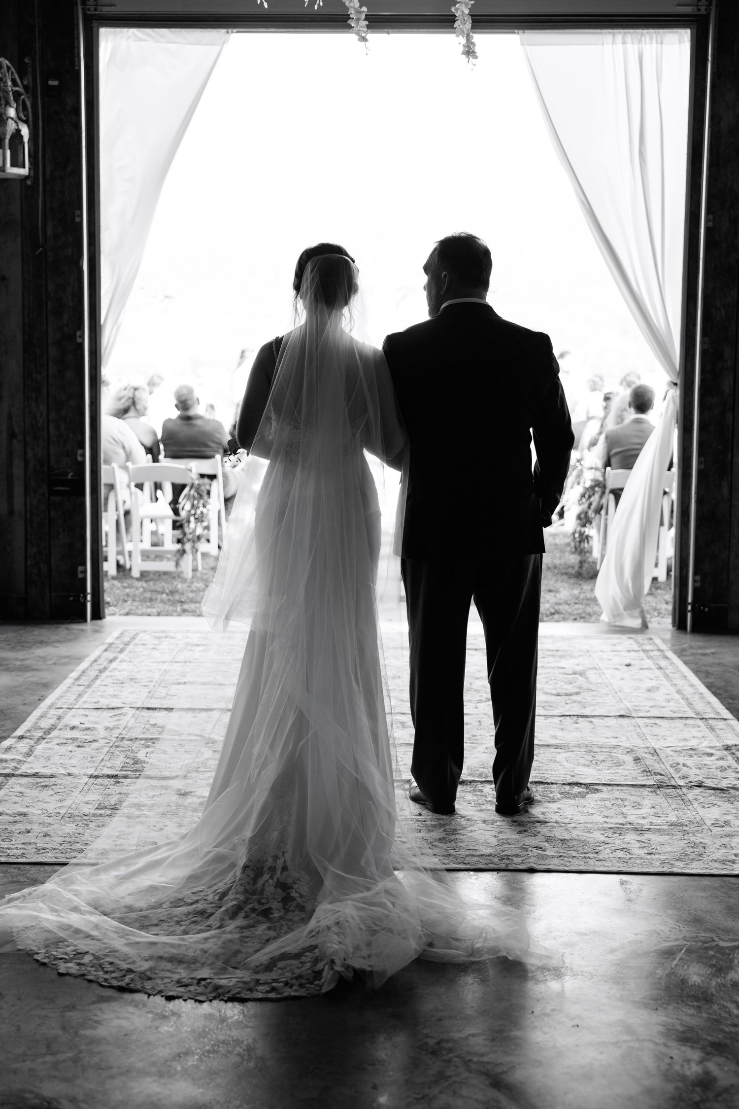 Bride and father stand in barn doorway before walking down the aisle at rustic wedding ceremony.