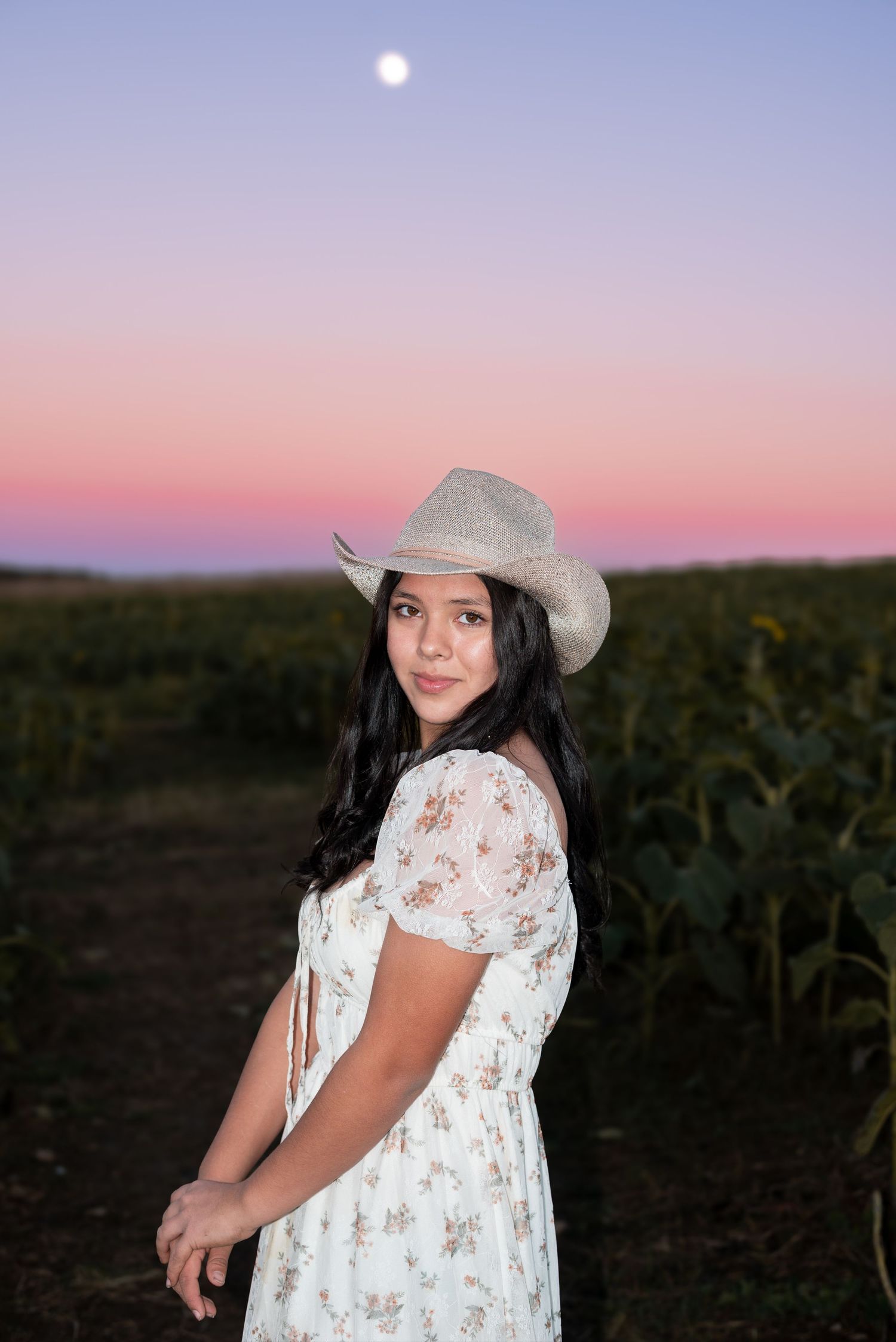 Person wearing cowboy hat and floral dress poses in field at sunset with pink sky.