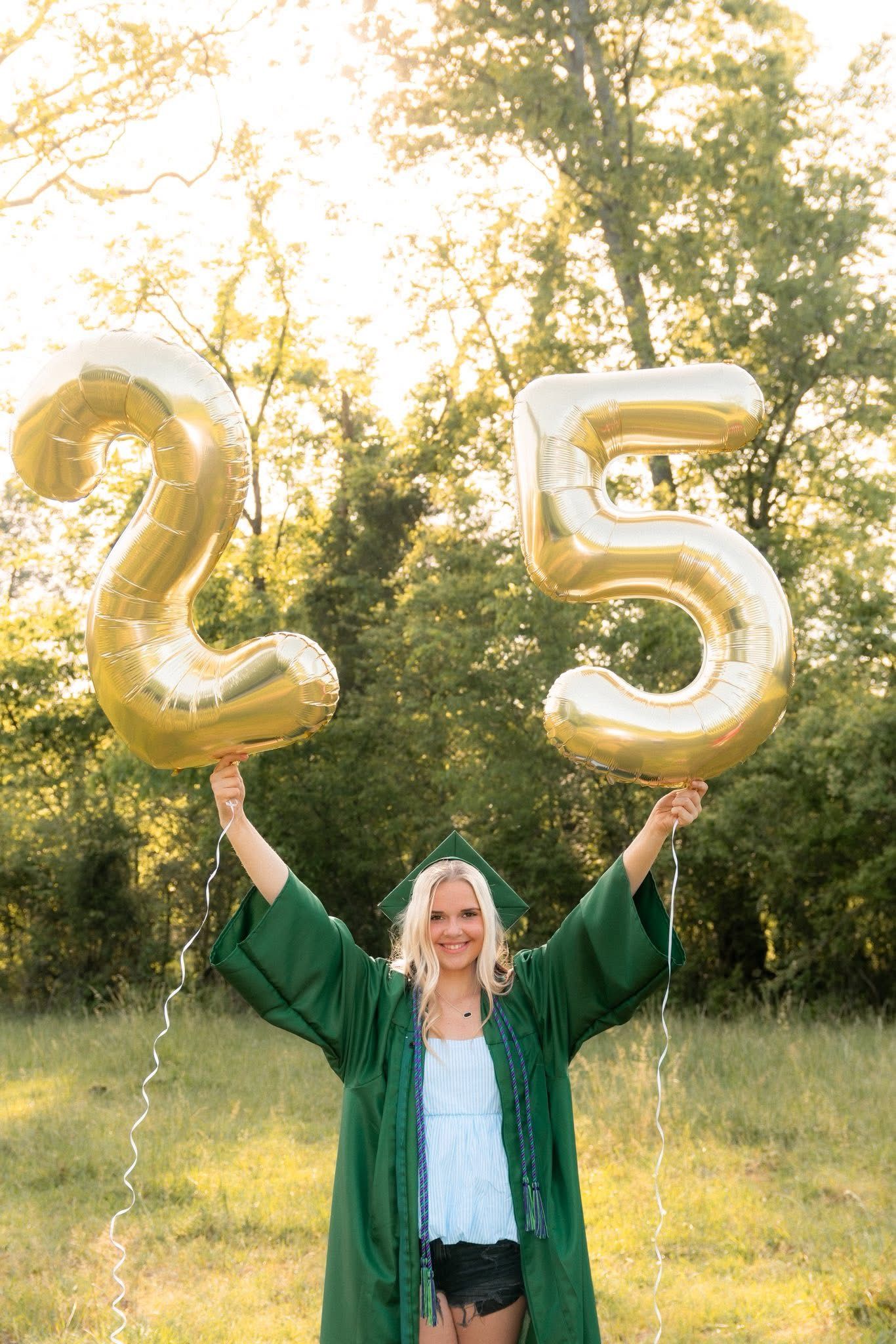 Graduate in green gown holding gold number 25 balloons in sunny field.