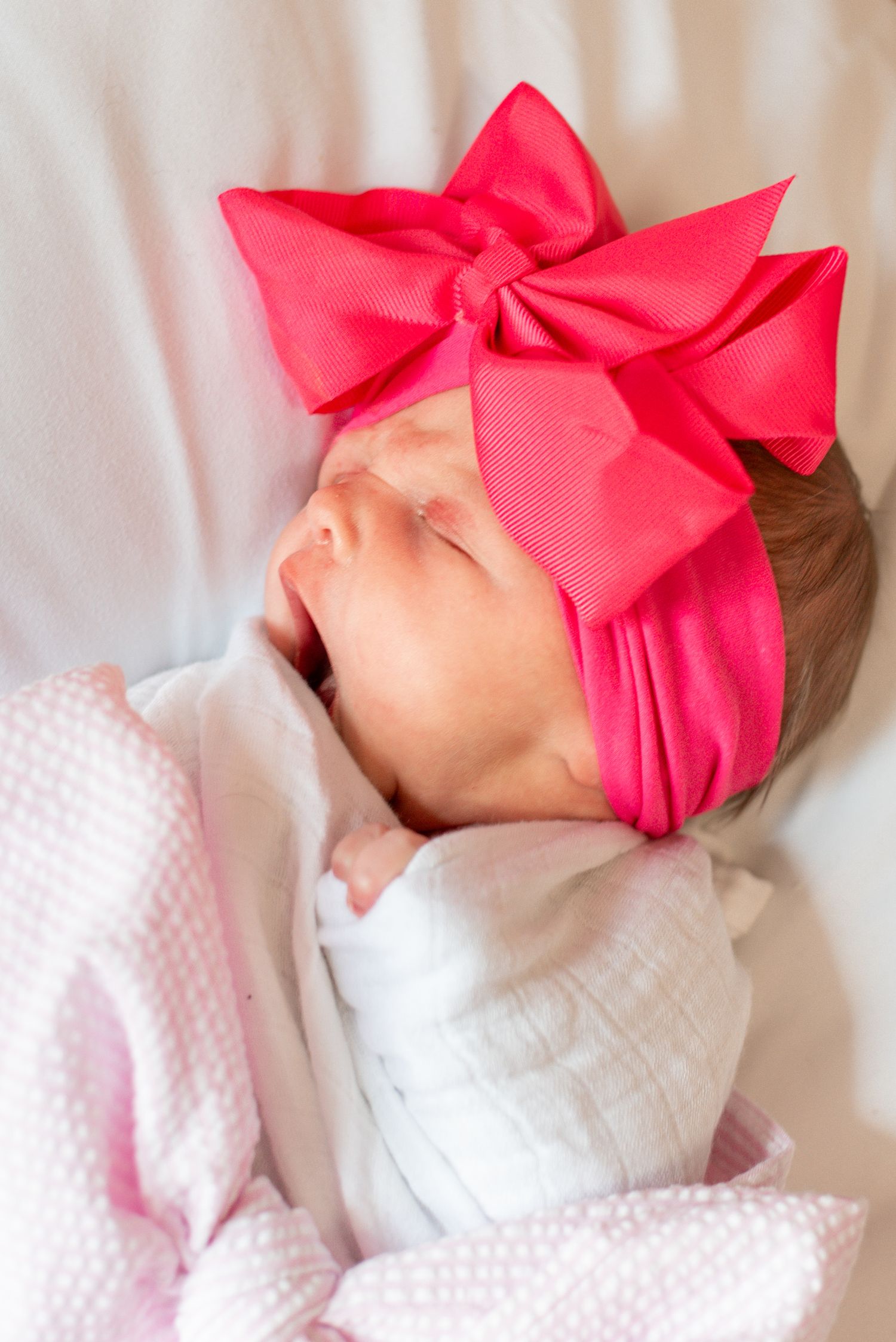 Sleeping newborn in white dress with large pink bow headband against white background.