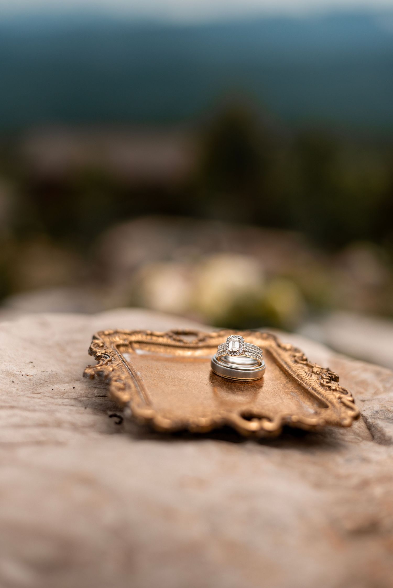 Wedding rings stacked on a ornate gold tray placed on a rustic stone surface with a blurred mountain backdrop.