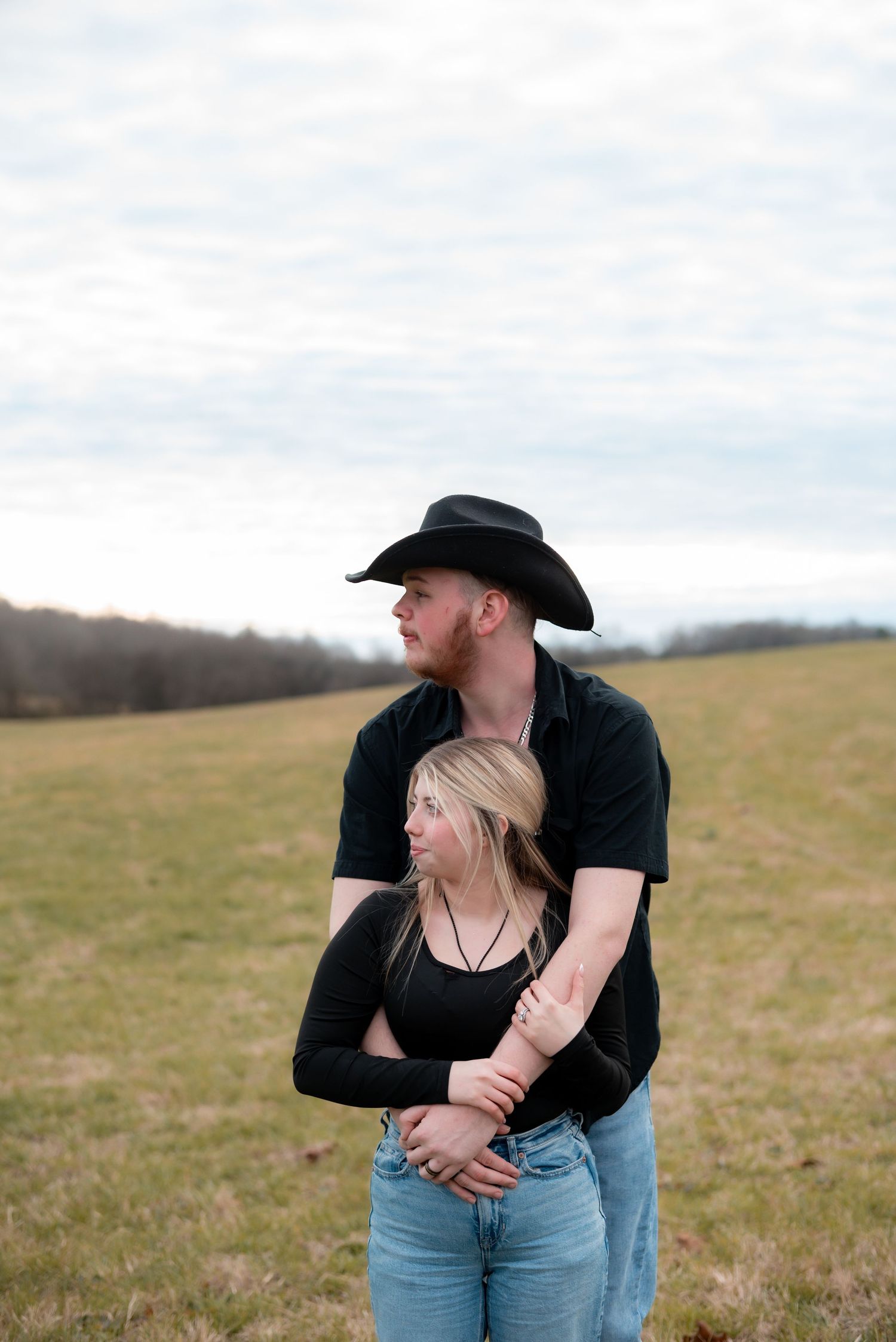 Couple embracing in casual attire on a grassy hillside under cloudy skies.