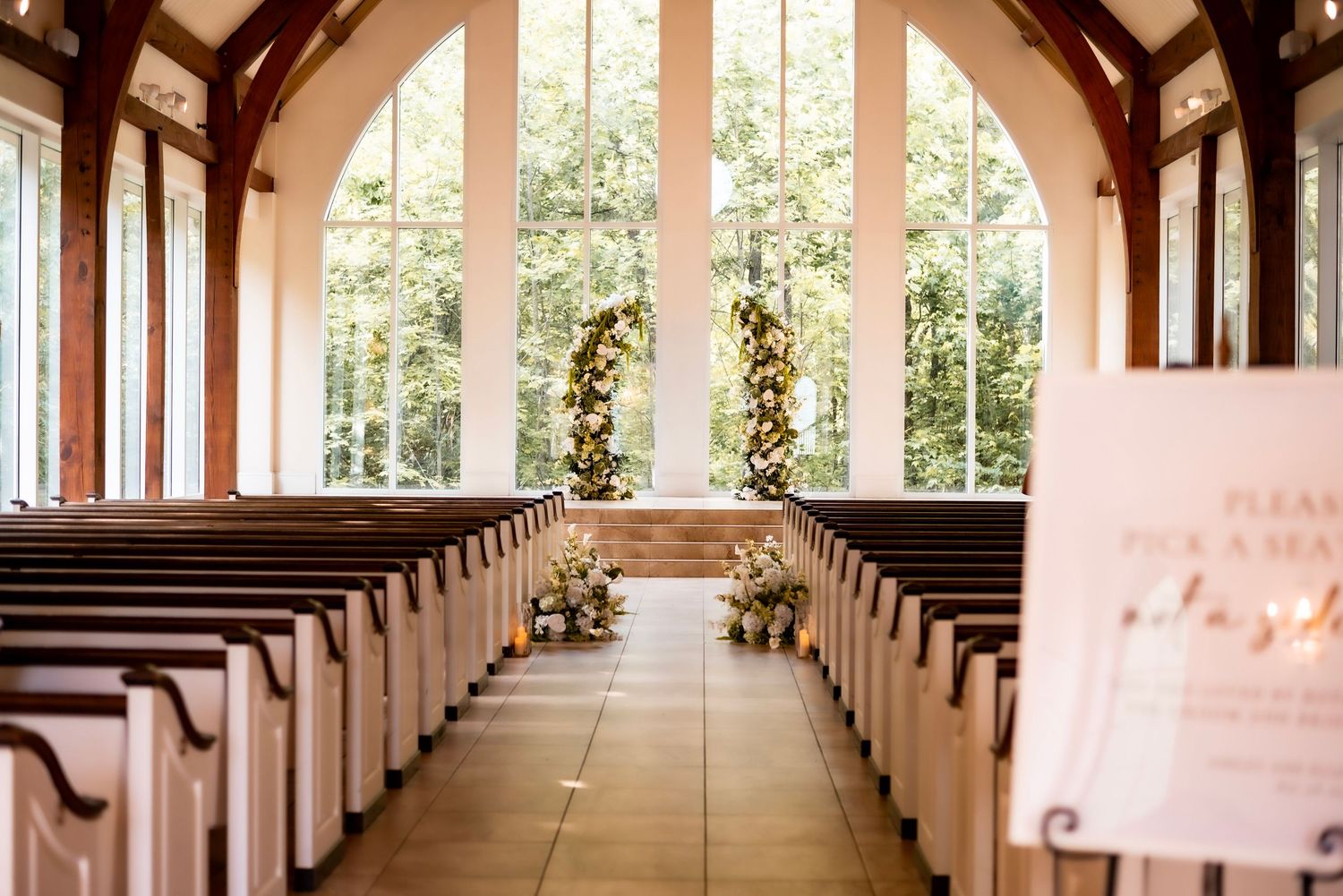 Elegant wedding chapel aisle with wooden pews, floral arch, and large arched windows overlooking lush greenery.