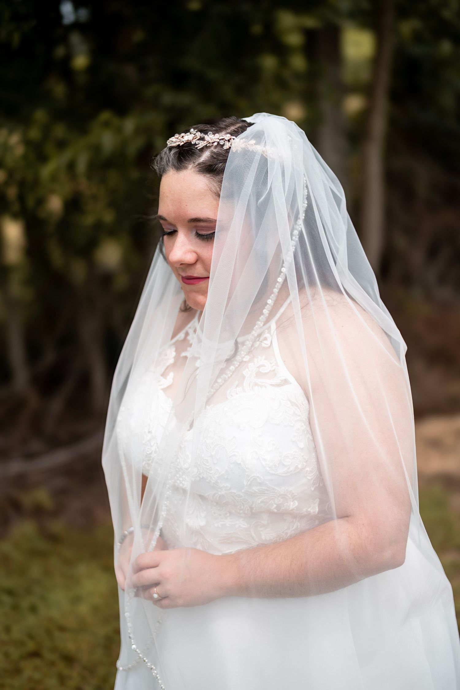 Bride in lace wedding dress and pearl-trimmed veil with floral headpiece, smiling softly outdoors.