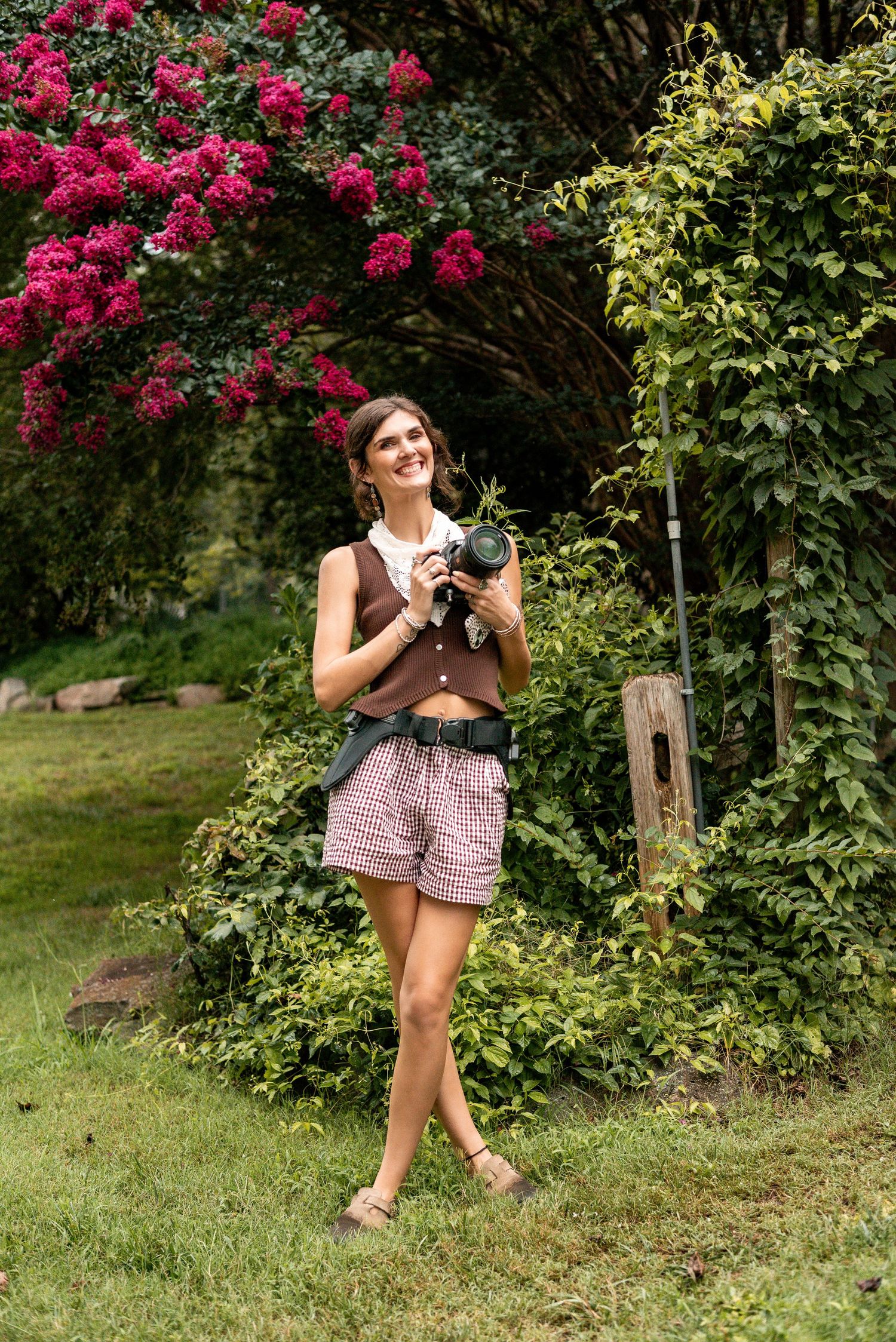 A smiling woman holds a camera in a lush garden with pink blooming flowers, wearing shorts and a brown vest.