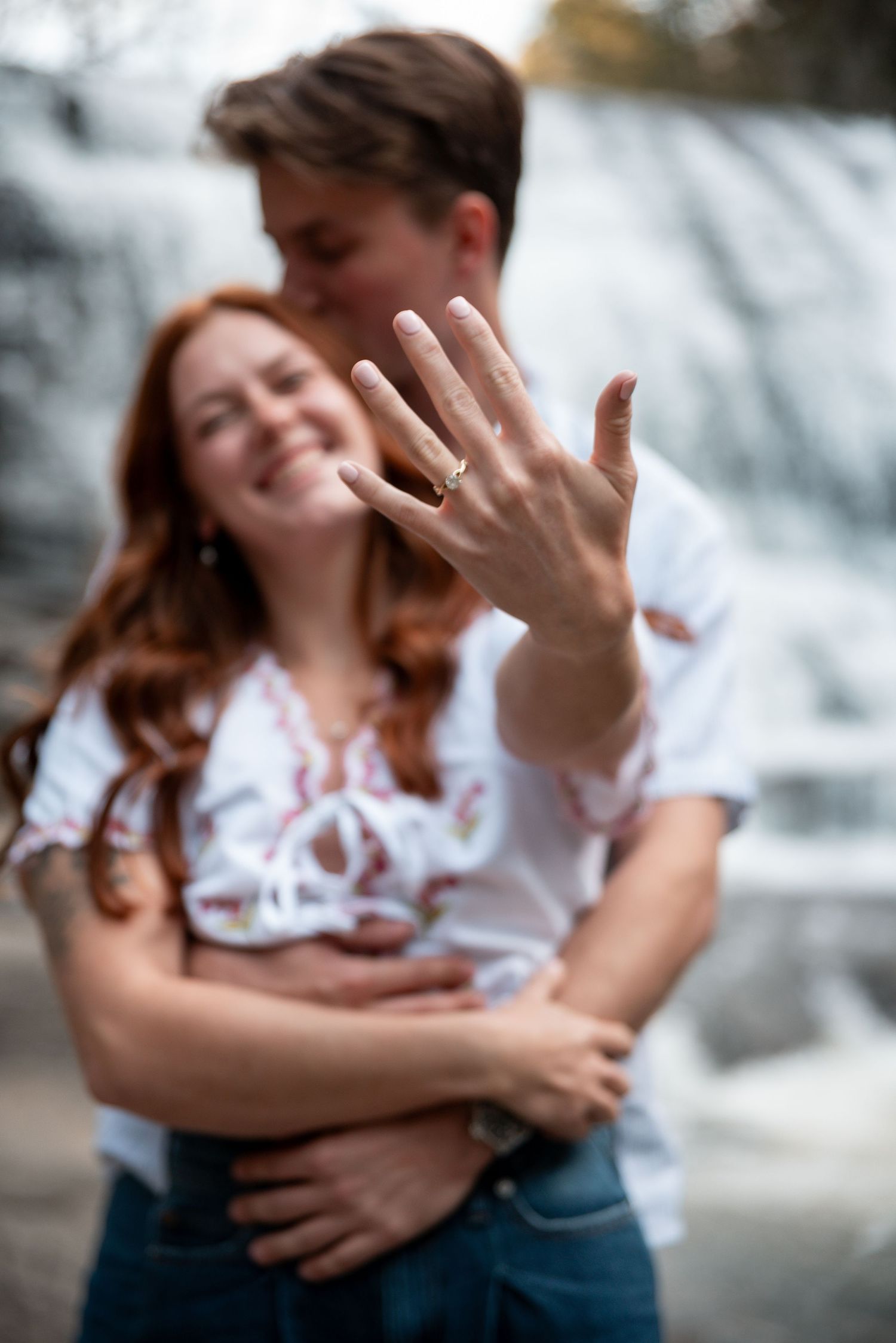 Happy couple showing off engagement ring in front of waterfall, woman with red hair smiling joyfully.