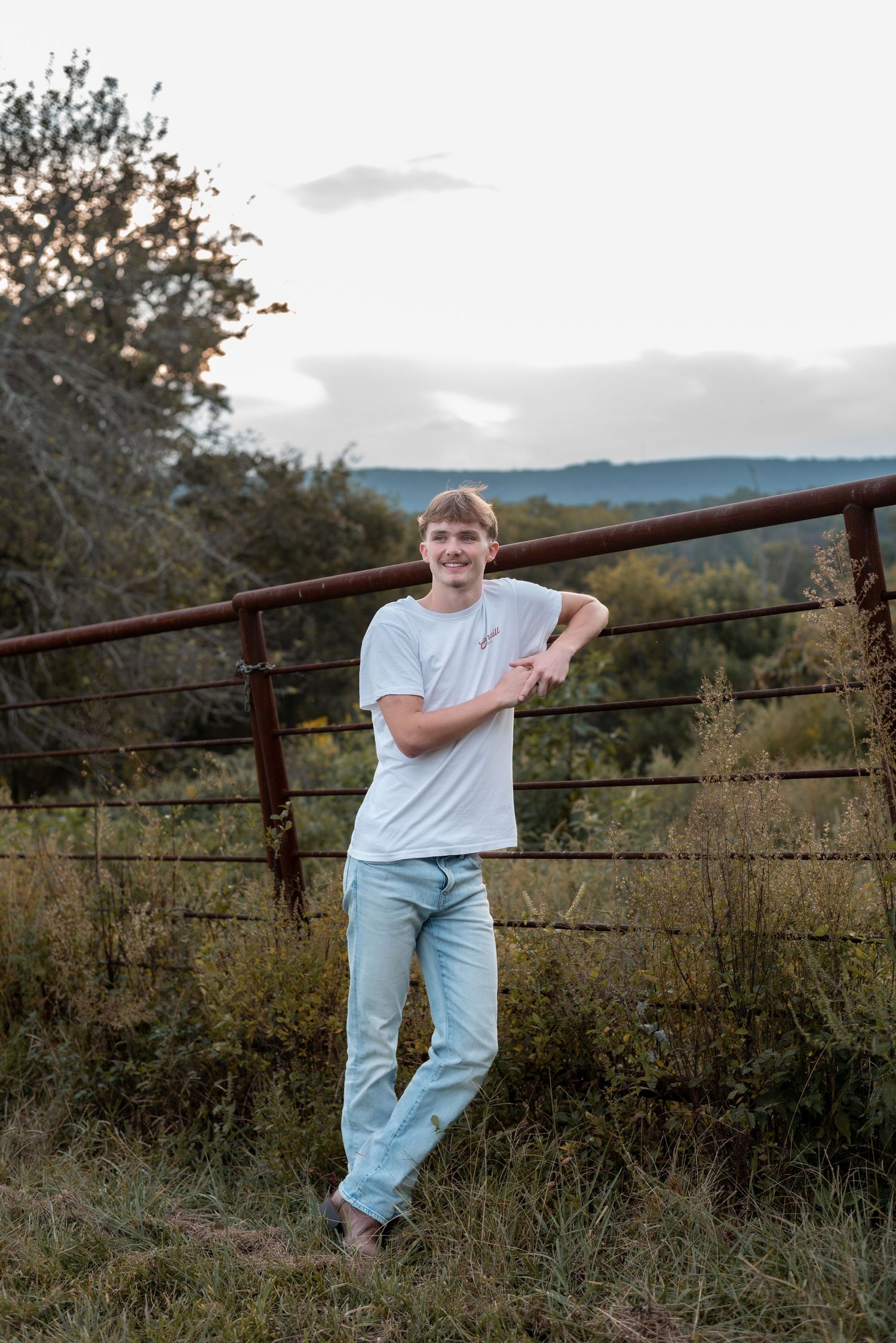 Person in light blue t-shirt leans against rustic metal fence at sunset.