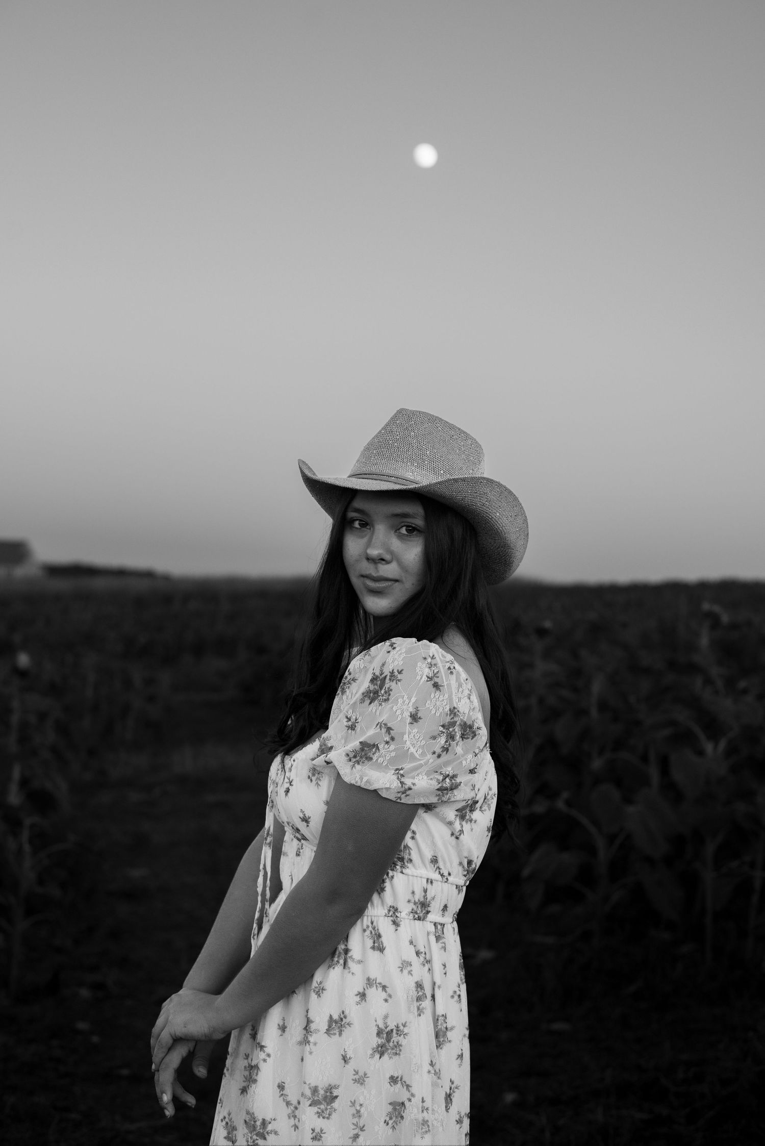 A person in a floral dress and cowboy hat poses against a twilight sky.