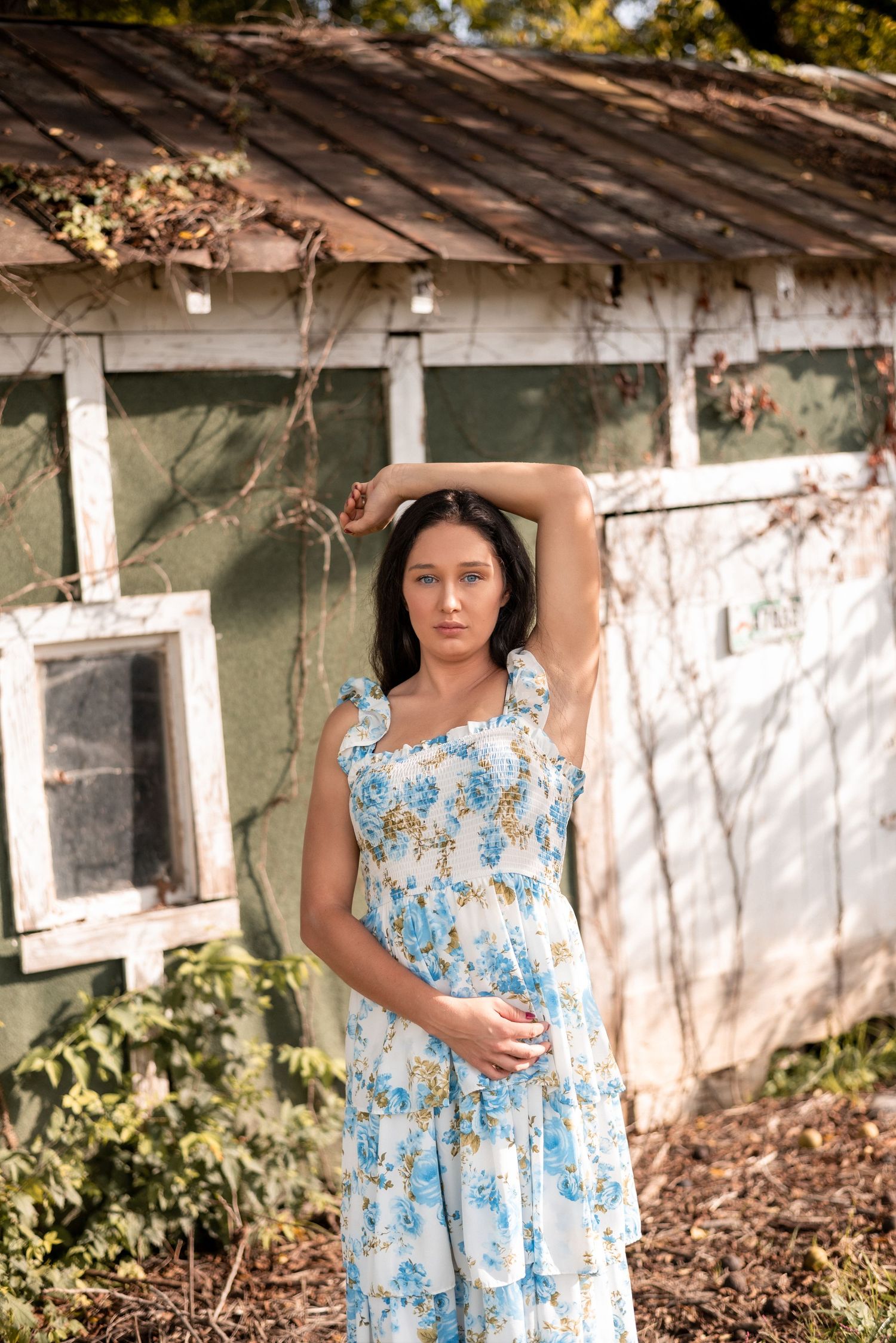 A person in a blue floral dress poses near a rustic building with peeling paint.