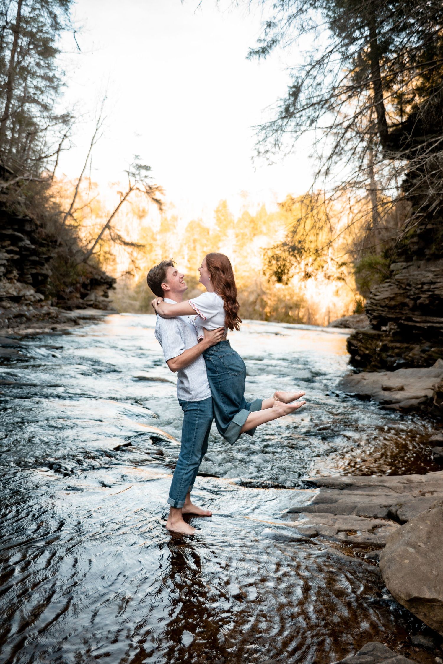 Man lifting woman in his arms while standing in a shallow river surrounded by golden sunlit trees.
