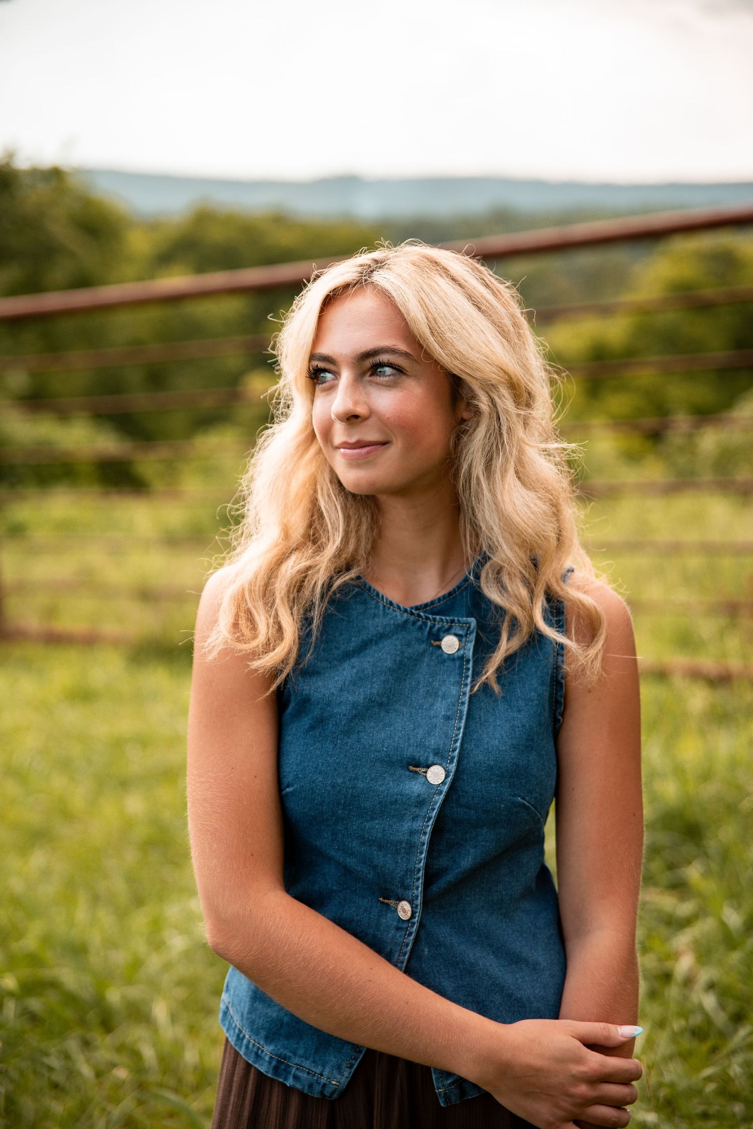 Young blonde woman in denim vest smiling outdoors in green field with rustic fence and hills backdrop.