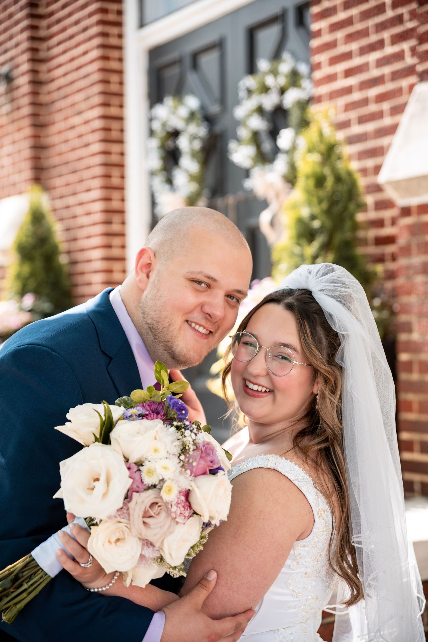 Smiling bride and groom pose with white and pink bouquet outside brick building on their wedding day.