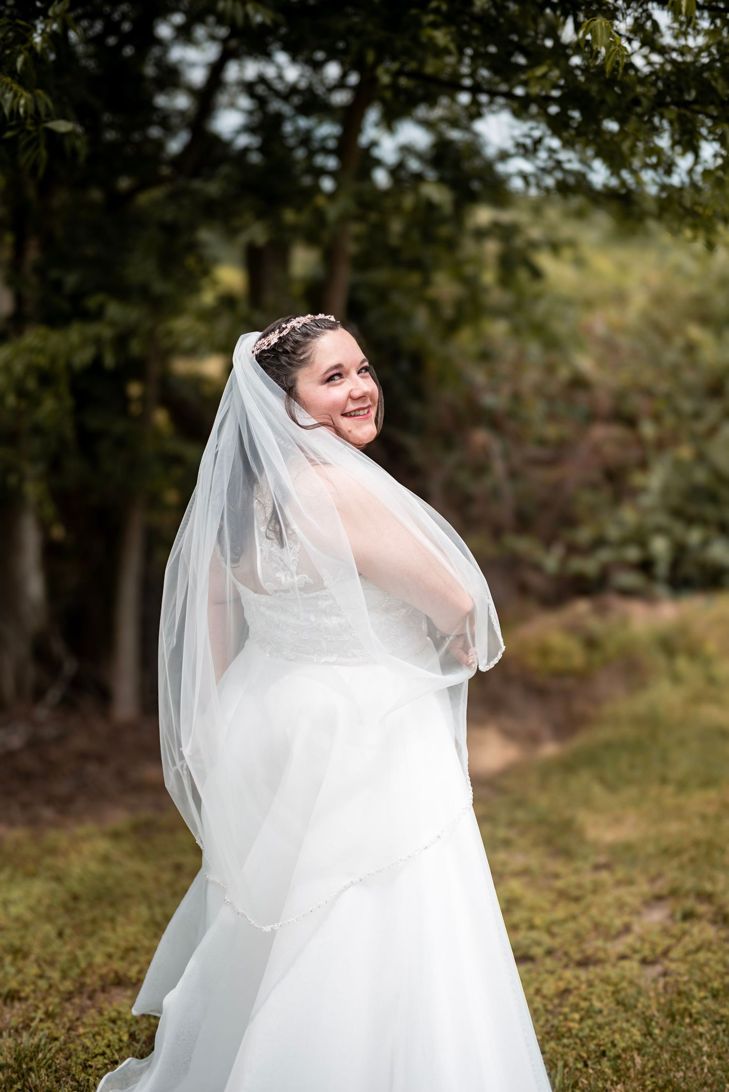 Smiling bride in white gown and veil poses outdoors among green trees on her wedding day.
