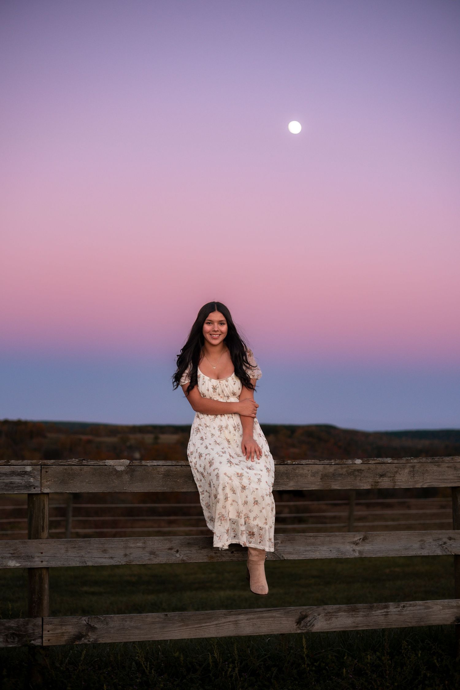 A person in a floral dress poses on a wooden fence at sunset with purple sky.