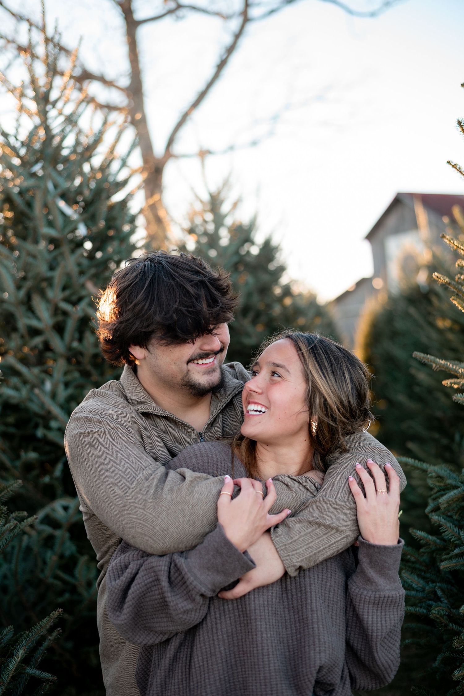 Couple embracing and laughing together among evergreen trees during a winter photoshoot.